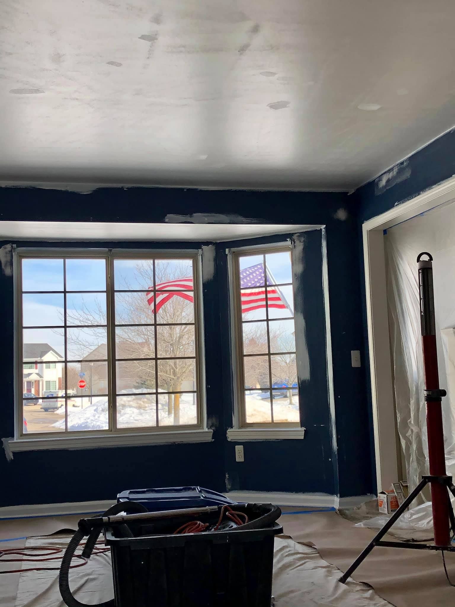 An indoor scene showing a room under renovation with dark blue walls, a white ceiling, and a bay window looking onto a flag.