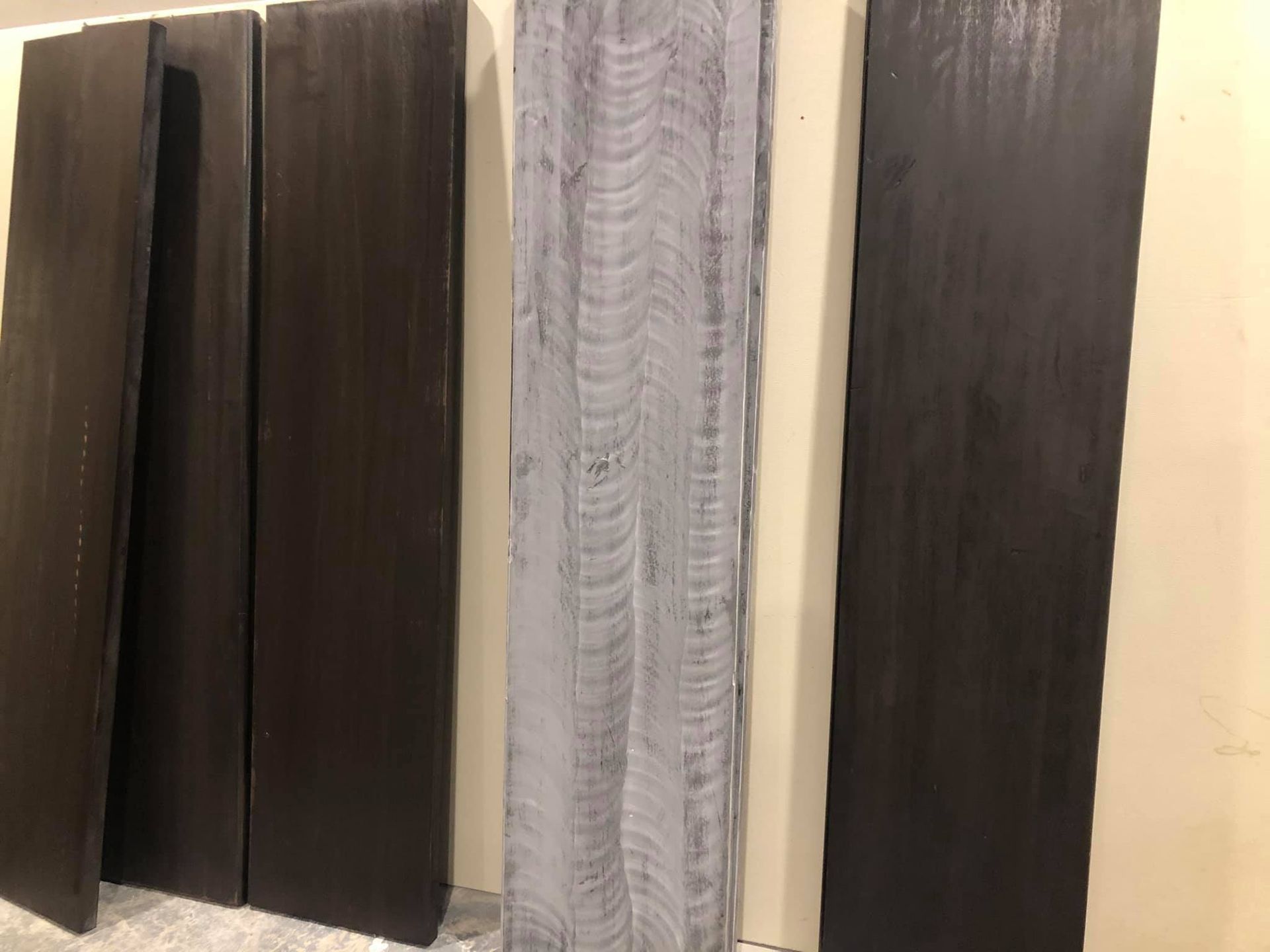 Five vertical wood planks leaning against a wall; four are stained dark brown, and one in the middle is light grey.