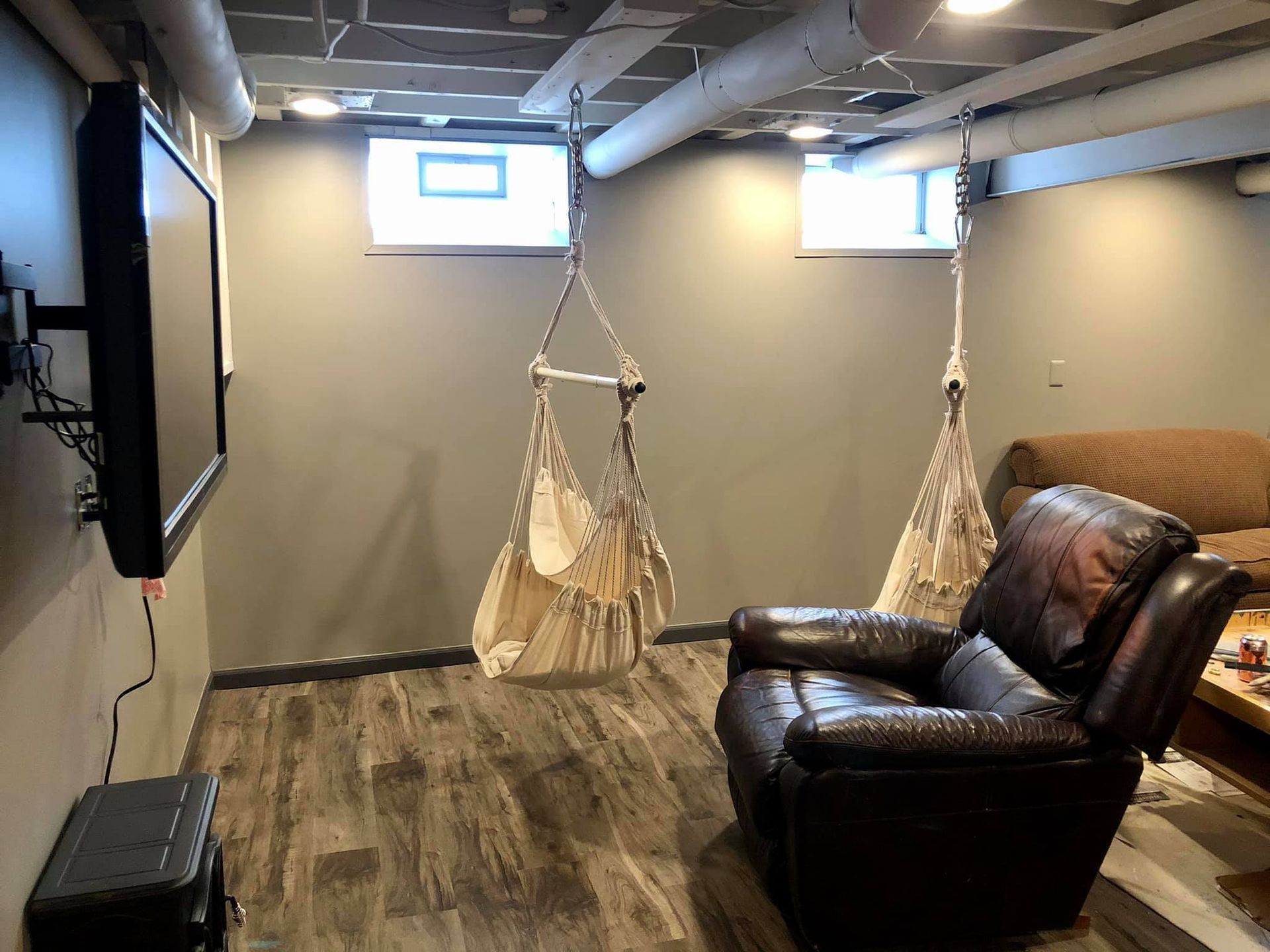 A basement lounge with two hanging canvas chairs, a dark leather recliner, and a wall-mounted TV under exposed pipes.