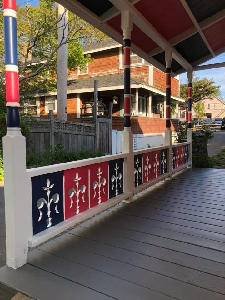 A porch railing with decorative panels featuring fleur-de-lis cutouts in alternating red and navy blue, under matching posts.