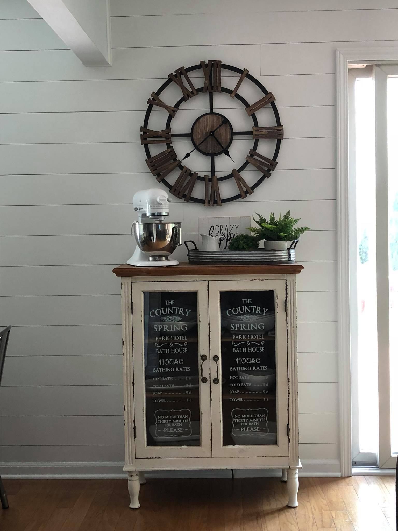 A white farmhouse-style cabinet with glass doors stands against a white shiplap wall, topped by a clock and mixer.