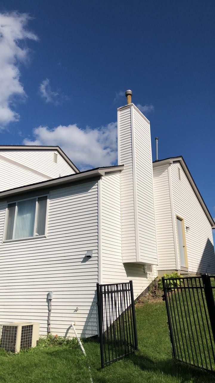 White house exterior with a prominent vertical chimney chase, siding, and a fenced yard under a blue sky.