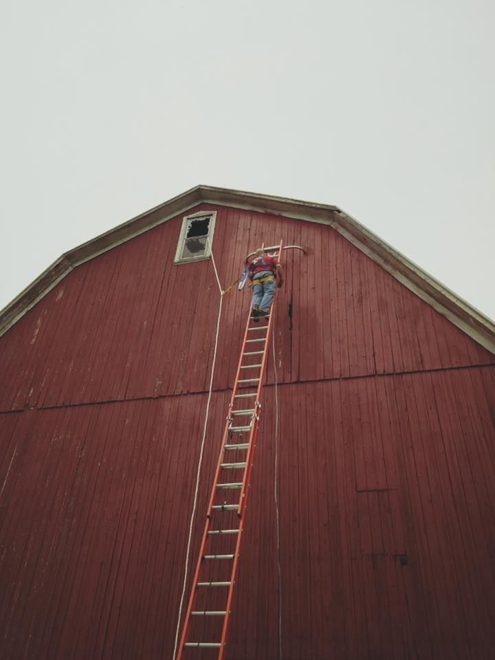A person on a tall ladder reaches toward a window on the side of a red barn.