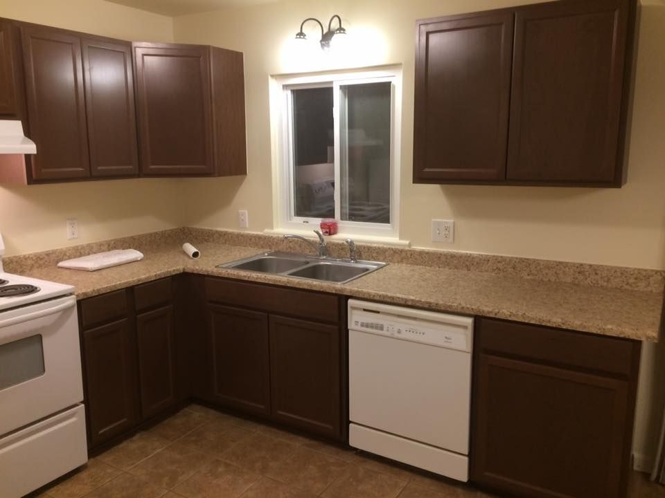 A kitchen with dark wood cabinets, beige countertops, a white oven, a white dishwasher, and a window above the sink.