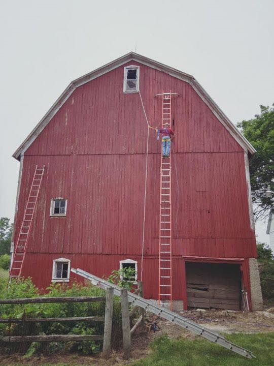 A red barn with a person standing on a tall ladder against the wall, reaching toward an upper-level window.