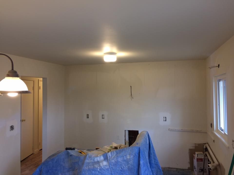 A room under renovation with light-colored walls, electrical outlets, a ceiling light, and a blue tarp covering furniture.
