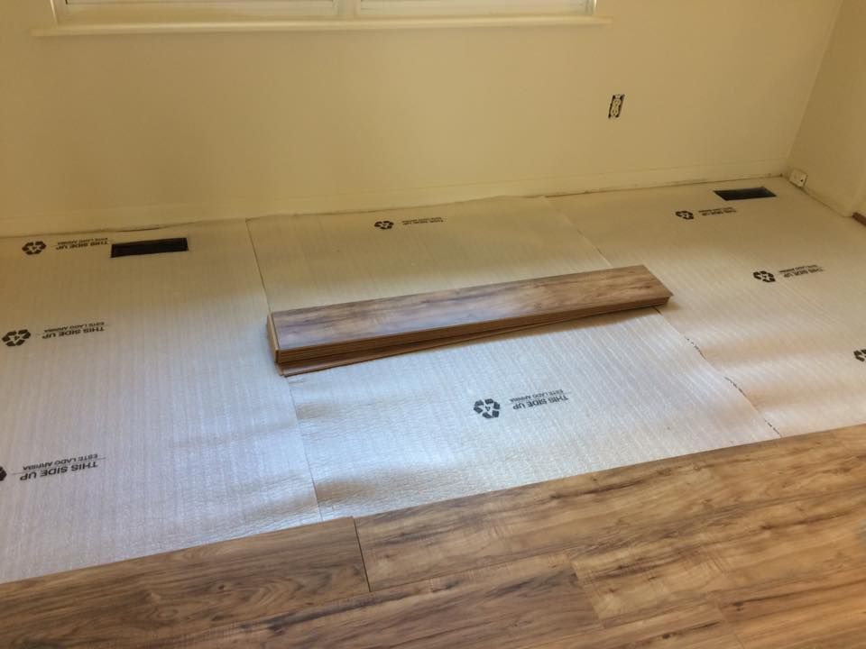 A room interior shows light-colored underlayment rolled out on the floor, partially covered by new wood-look plank flooring.