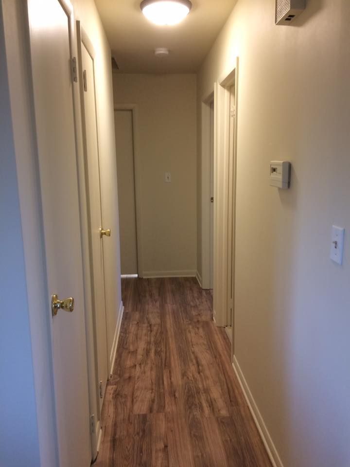 A brightly lit hallway with light beige walls and wood-patterned flooring, featuring several closed doors along both sides.