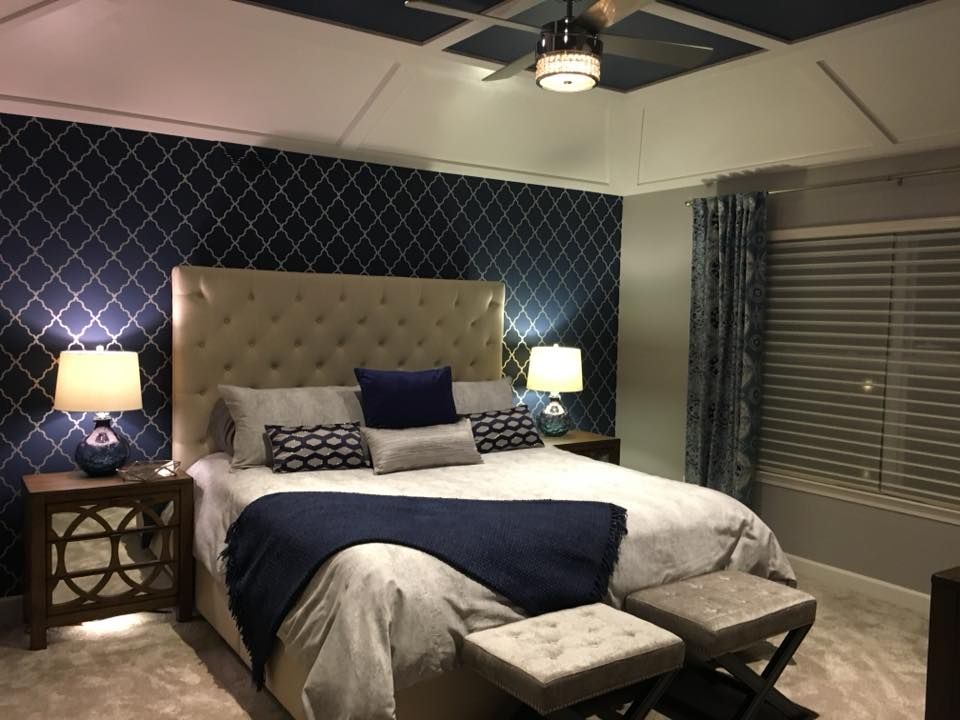 A luxurious bedroom with a tufted beige headboard, navy lattice wallpaper, a tray ceiling, and two upholstered ottomans.