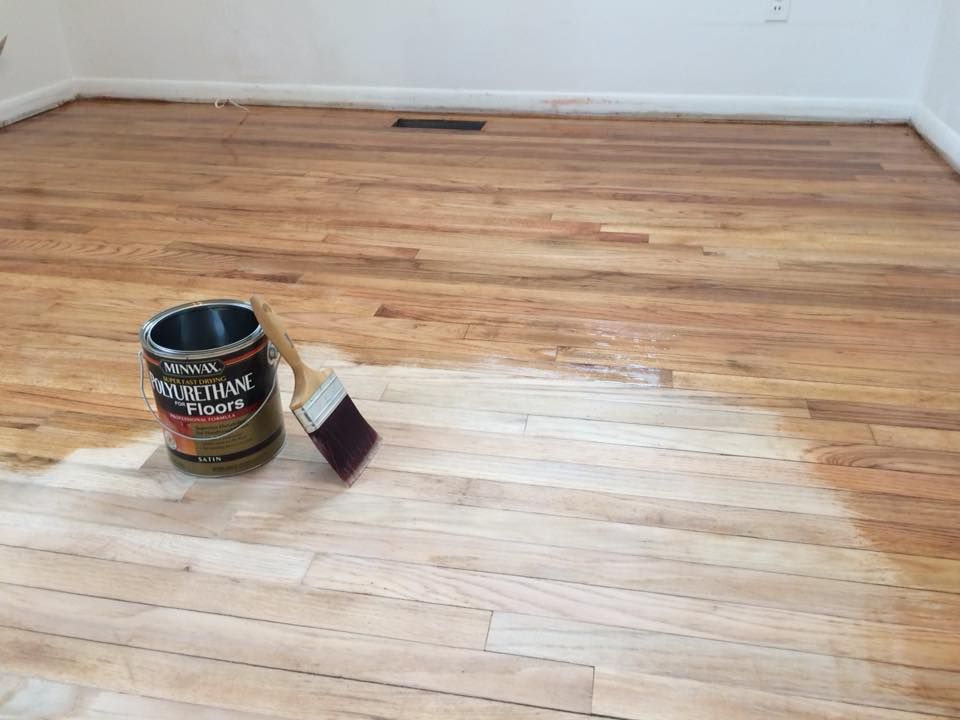 A can of wood floor polyurethane and a brush sit on a hardwood floor, showing a half-finished area of light raw wood.