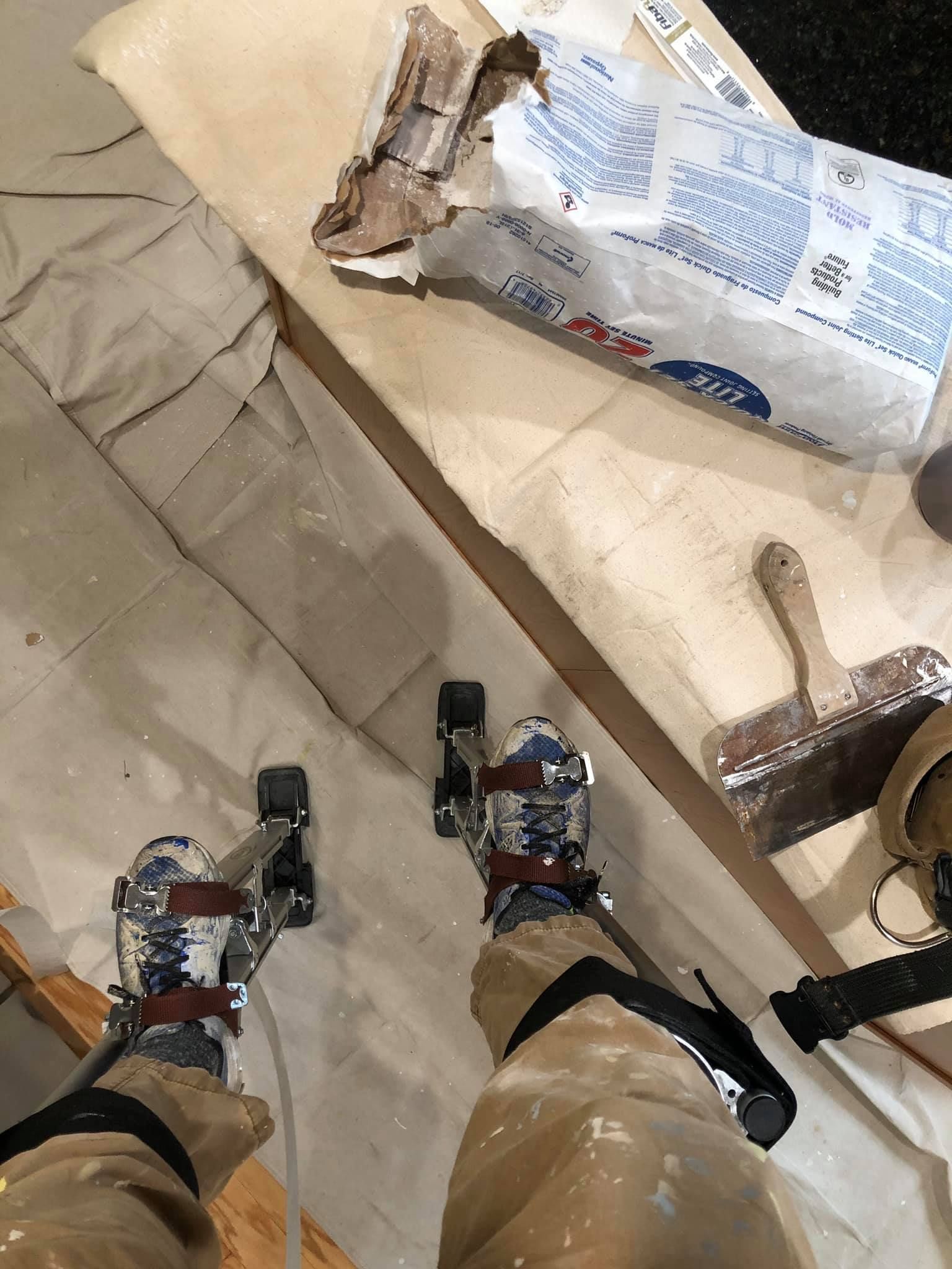 A person wearing drywall stilts stands on a drop cloth next to a bag of joint compound and a taping knife.