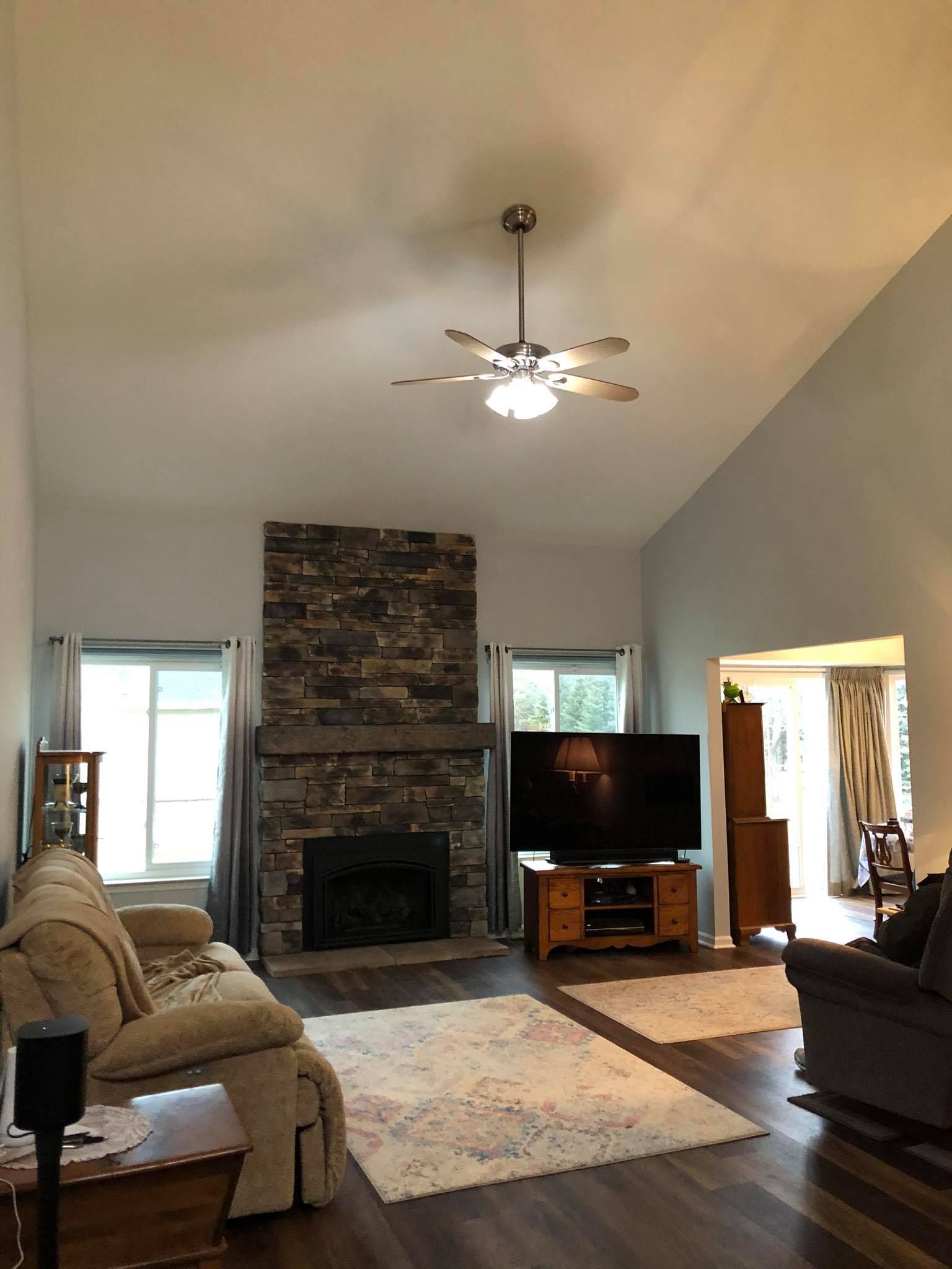 A living room with a stone fireplace, a TV, wood-look flooring, area rugs, a ceiling fan, and light-colored furniture.