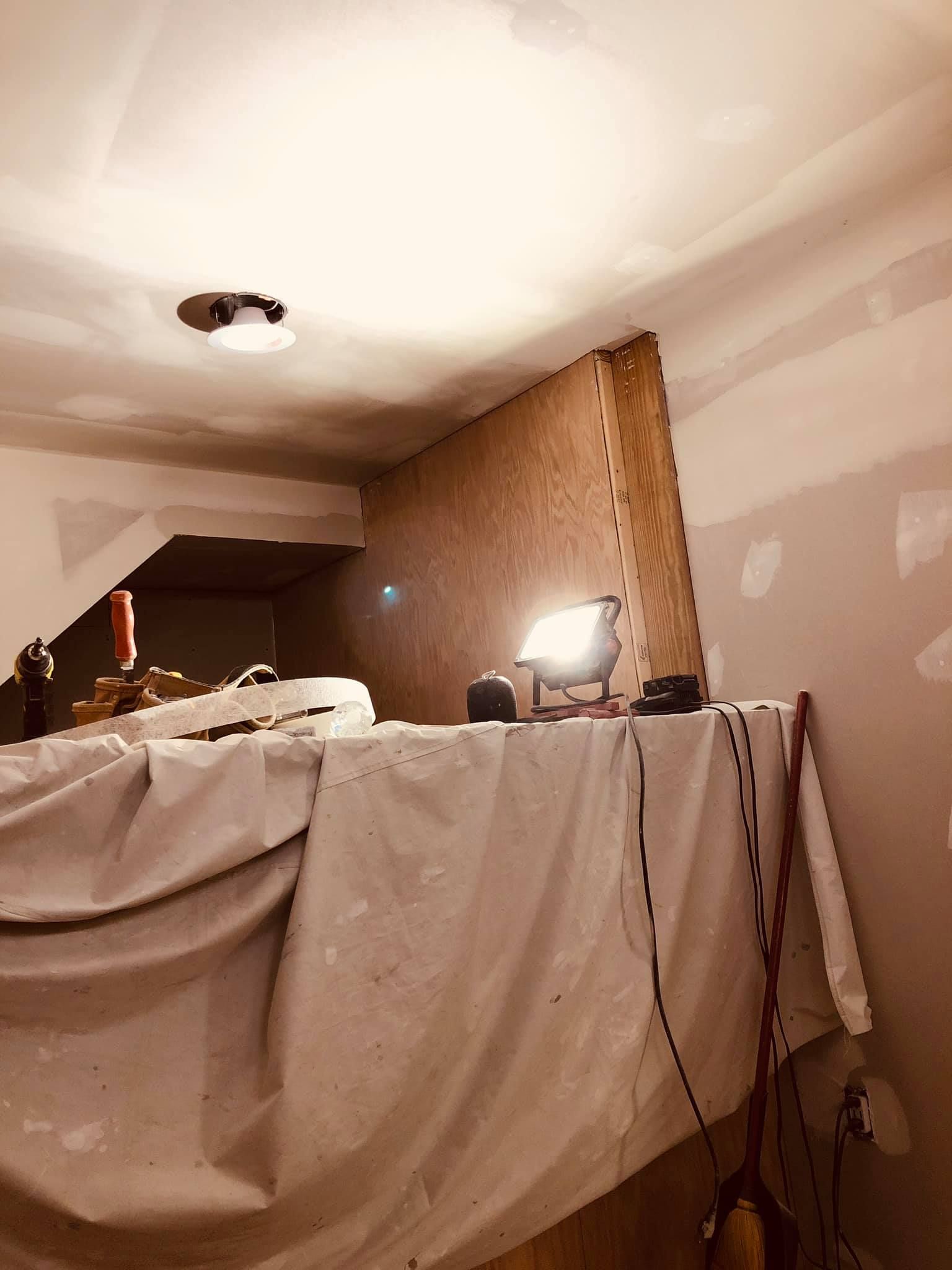 A renovation scene featuring a wooden wall structure, a bright portable work light, and a draped drop cloth.