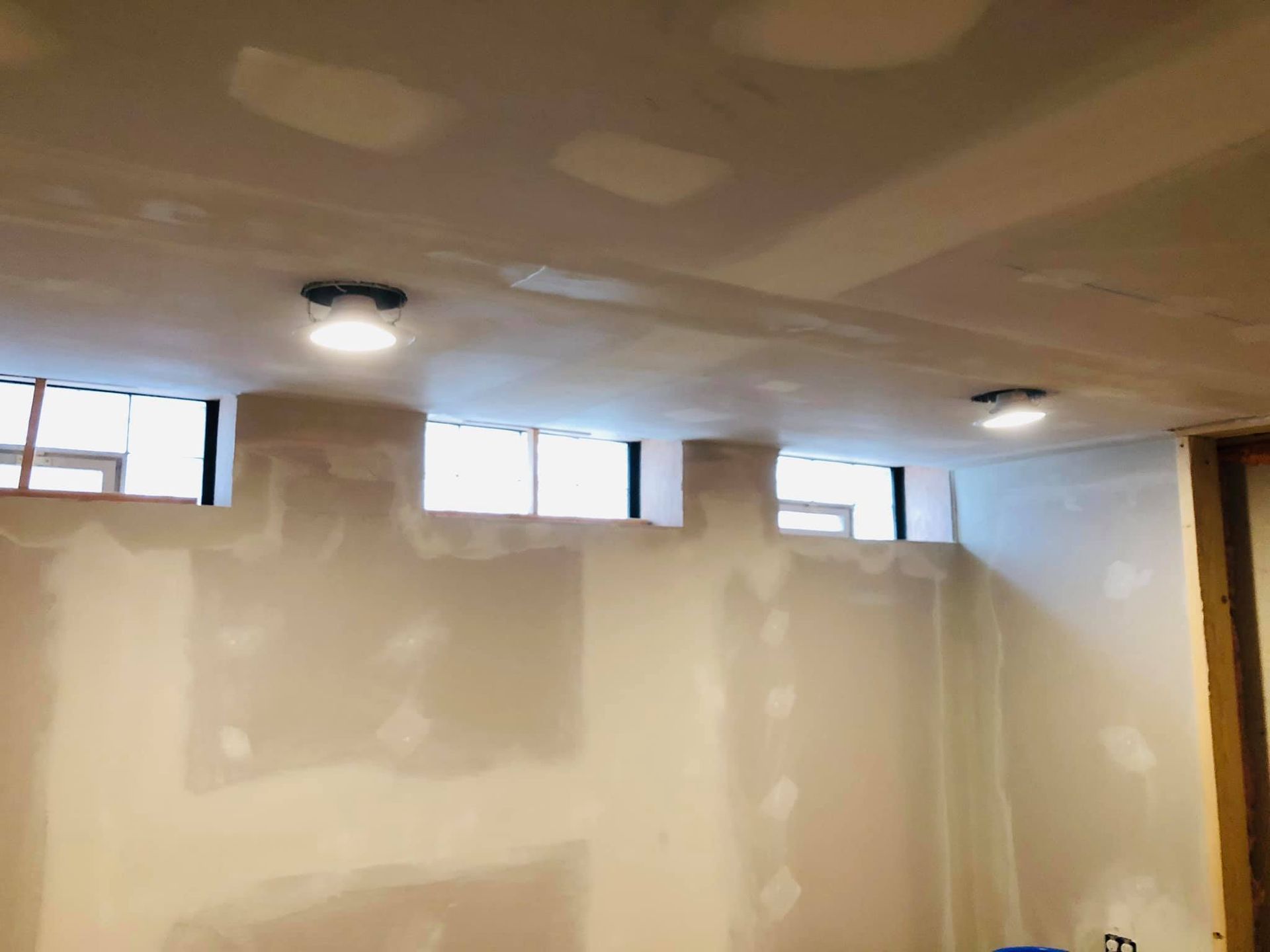 An unfinished basement room with drywall on walls and ceiling, including patches of spackle and three recessed lights.