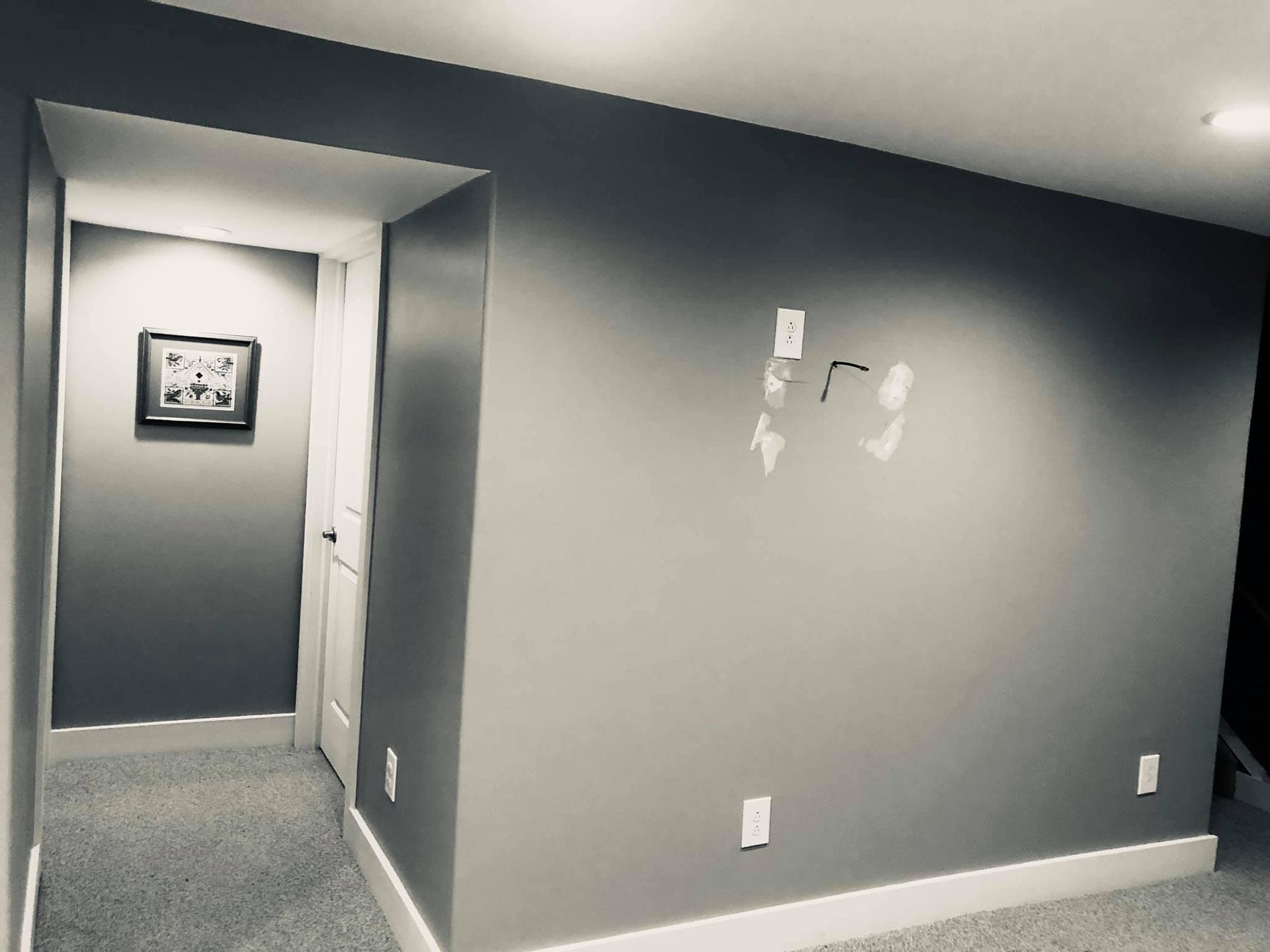 A gray room with carpeted floor, a hallway leading to a door with a framed picture, and a wall with small repair patches.