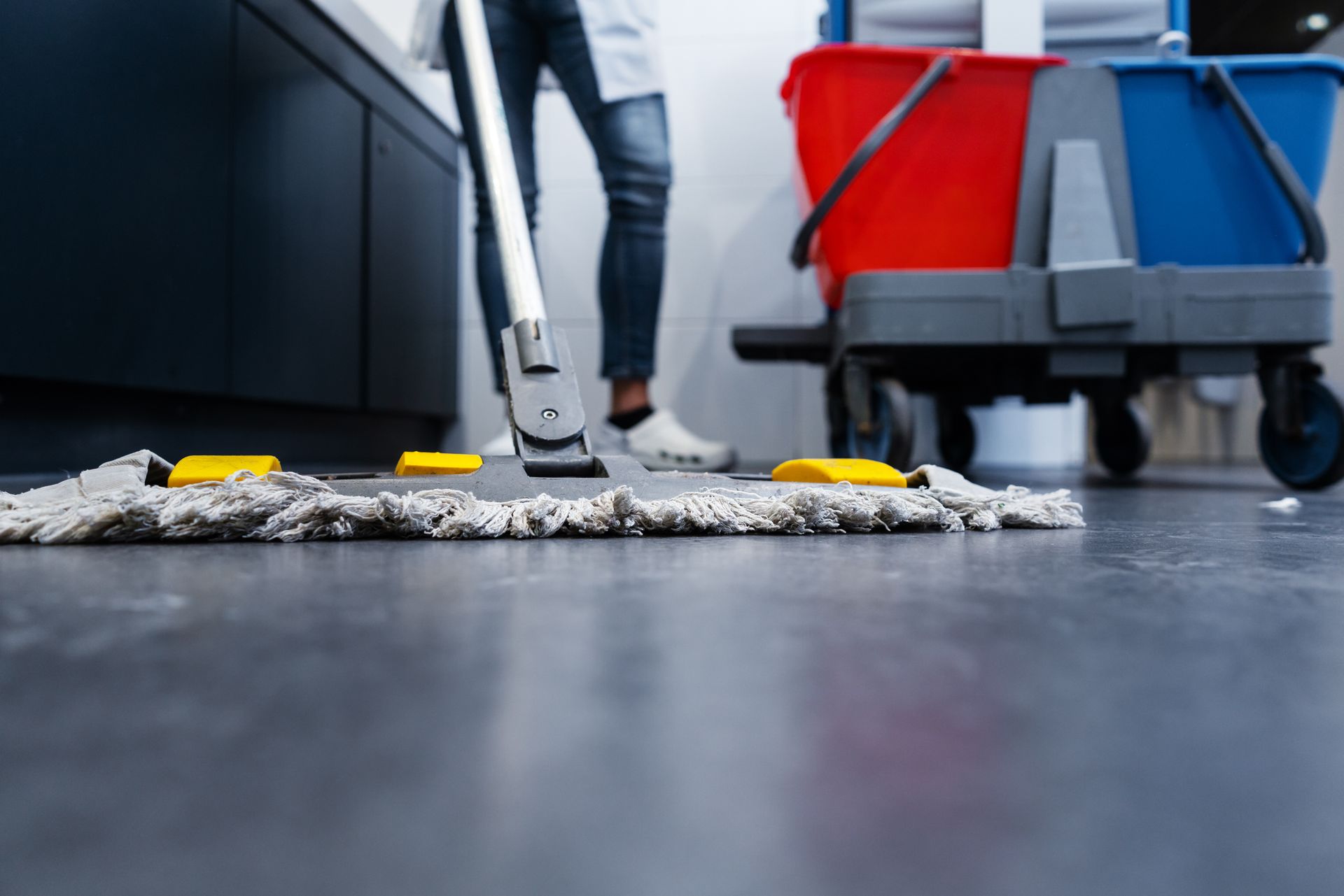 Close-up view of a janitorial worker using a mop to clean the restroom floor, with her cleaning trolley positioned nearby.