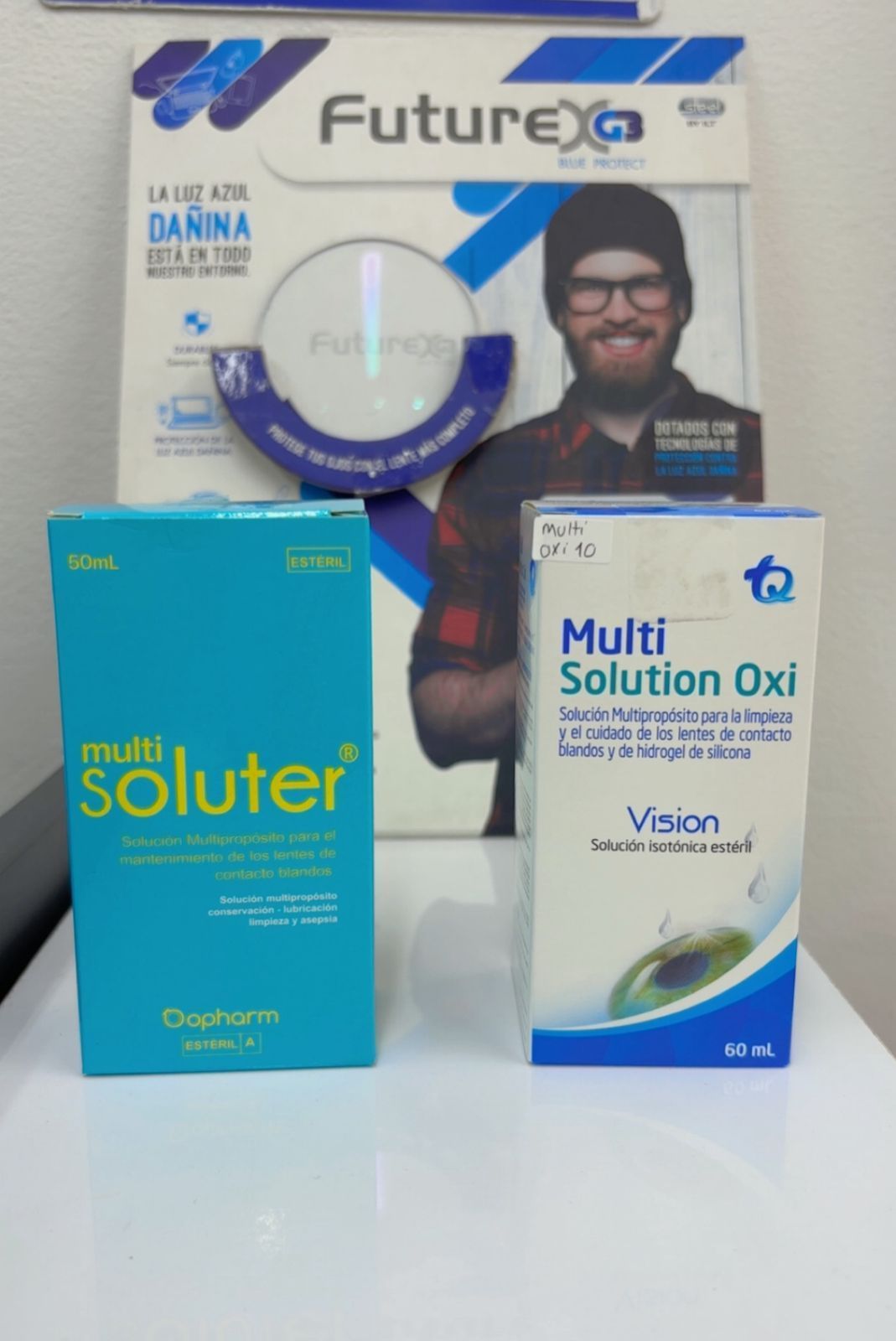 Dos cajas de solución oftálmica junto a una revista con un hombre. Cajas verde azulado y blancas sobre una superficie blanca.