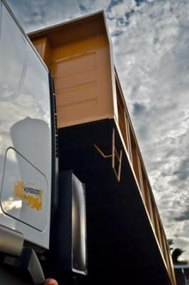 Caja de camioneta amarilla sobre un marco negro, unida a una camioneta blanca con el logotipo de una empresa, cielo nublado.