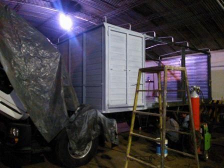 Caja de camión con paneles pintados de color gris, escalera, equipo de soldadura y lona en un taller.