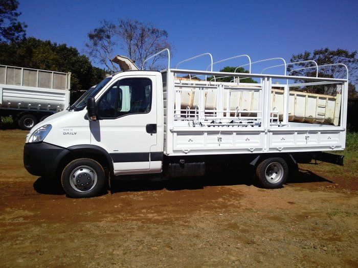Camión blanco con caja de carga enrejada estacionada sobre tierra frente a árboles.