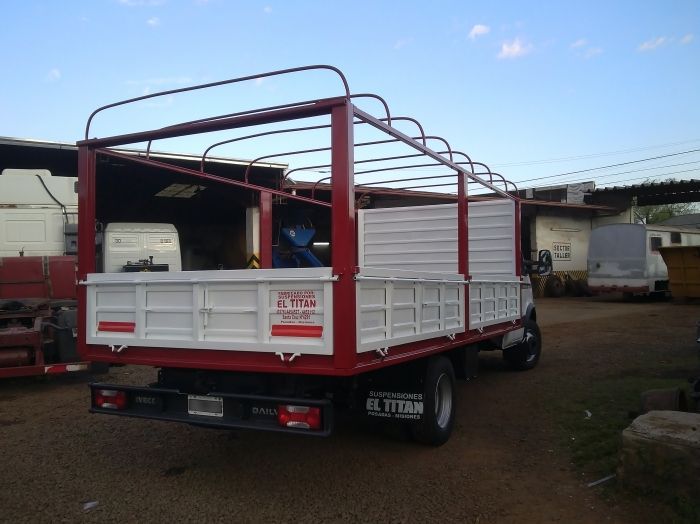 Camión de carga rojo y blanco, estructura de metal, laterales abiertos, estacionado en el exterior, día soleado.