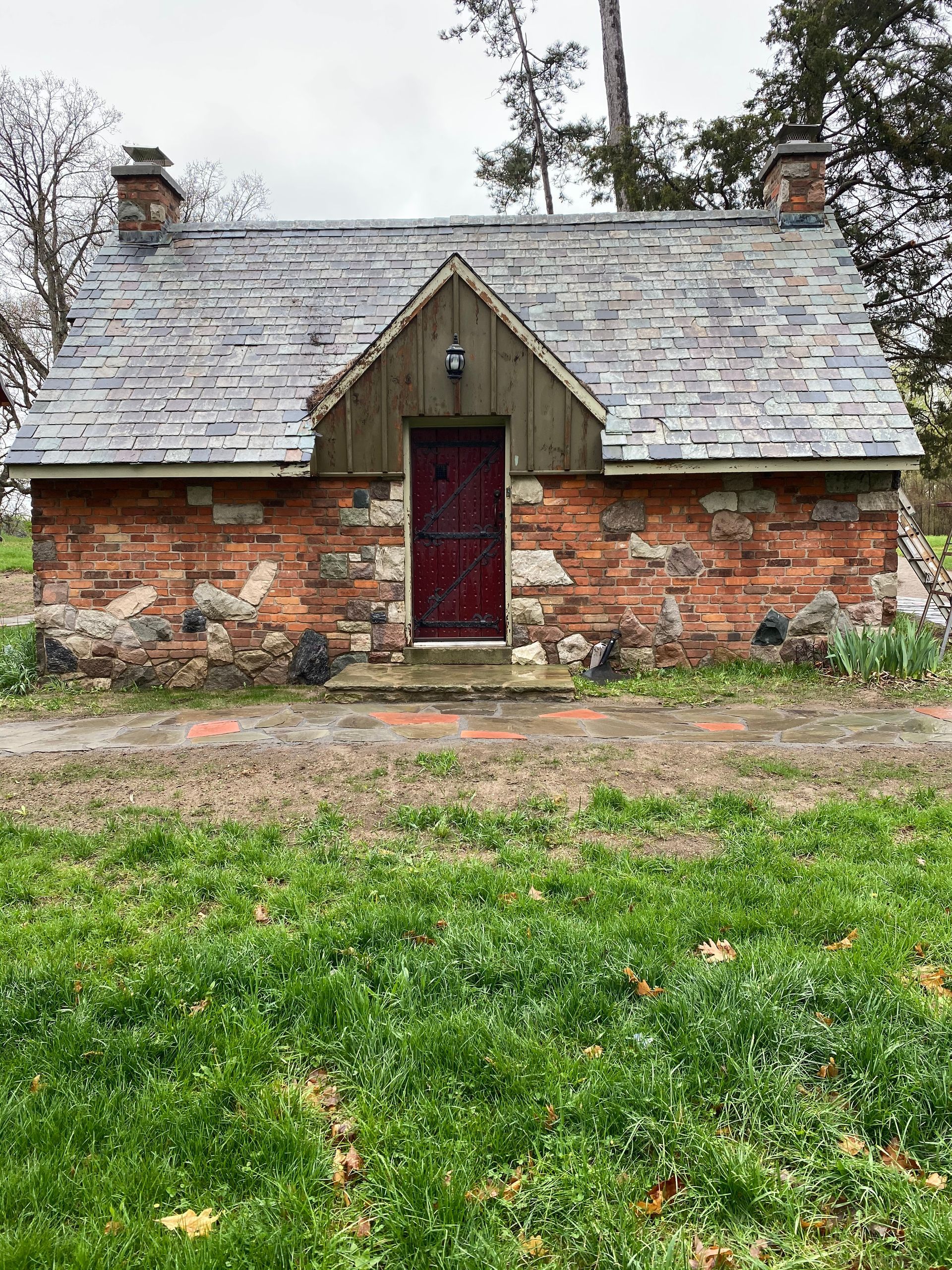A small brick house with a slate roof and a red door is sitting in the middle of a grassy field.