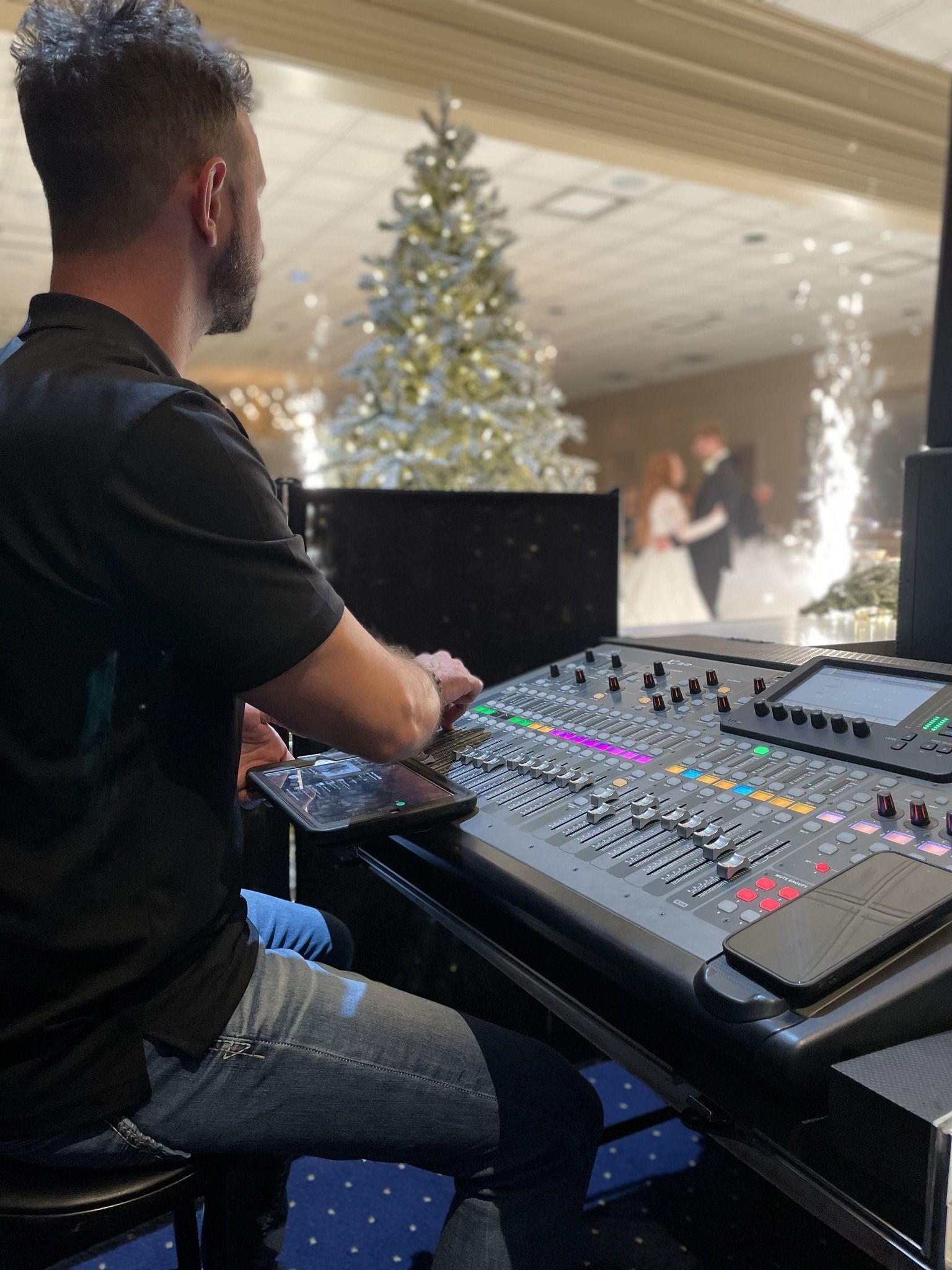 A person works at a large sound mixing console at a wedding with a Christmas tree and pyrotechnics in the background.