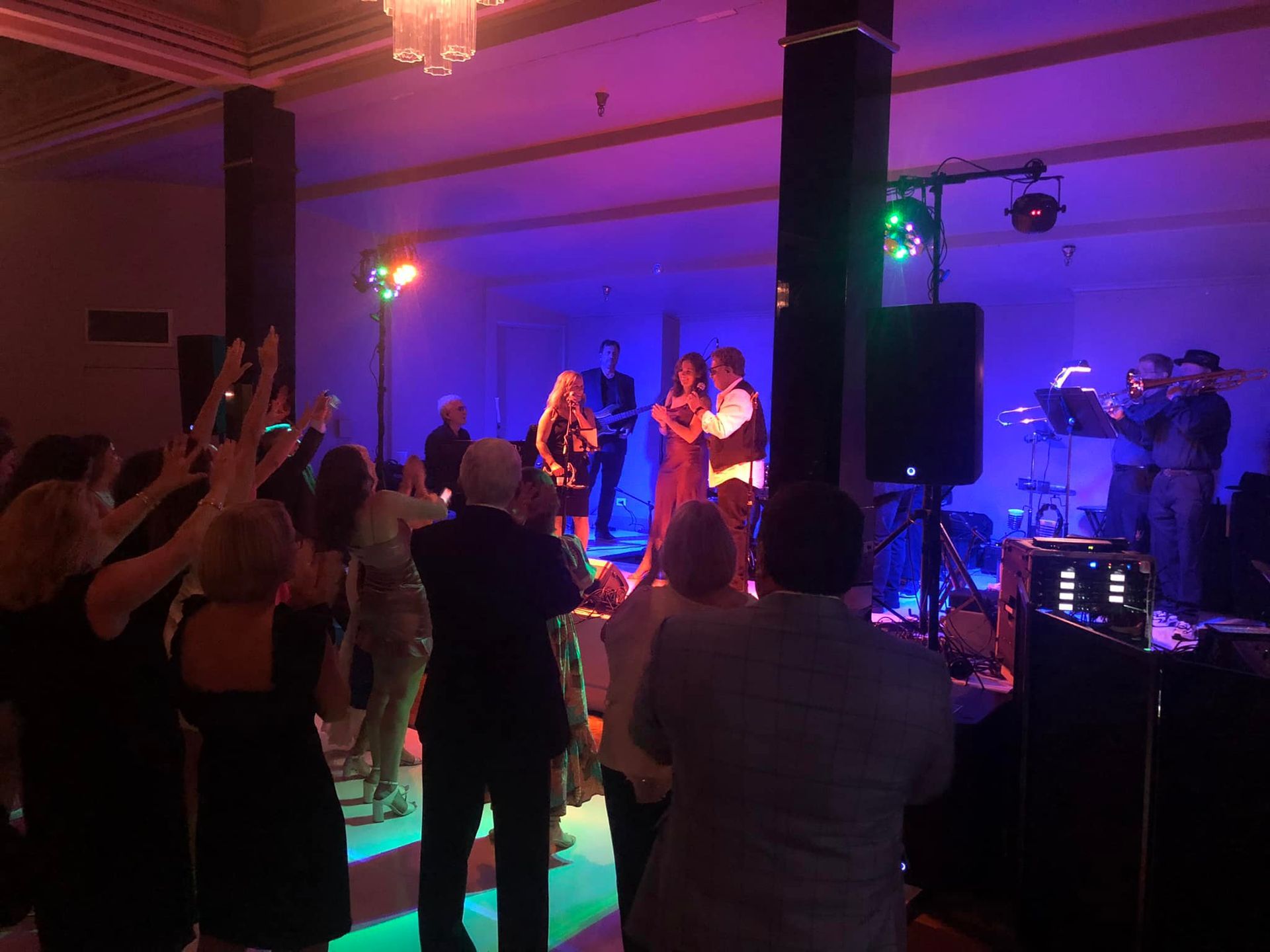 People enjoy a performance in a dimly lit event space illuminated with vibrant purple and green stage lights.