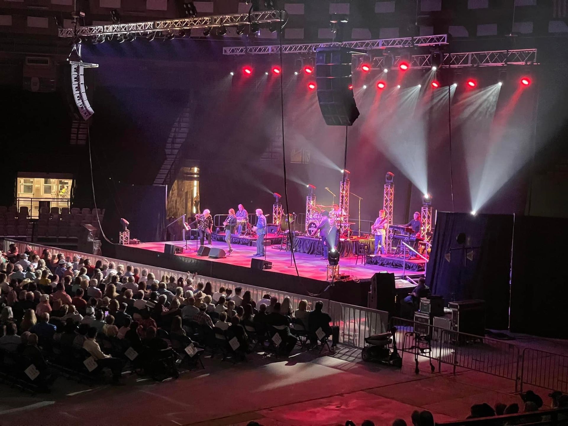 A band performs on a brightly lit concert stage before a large, seated audience in a darkened arena.
