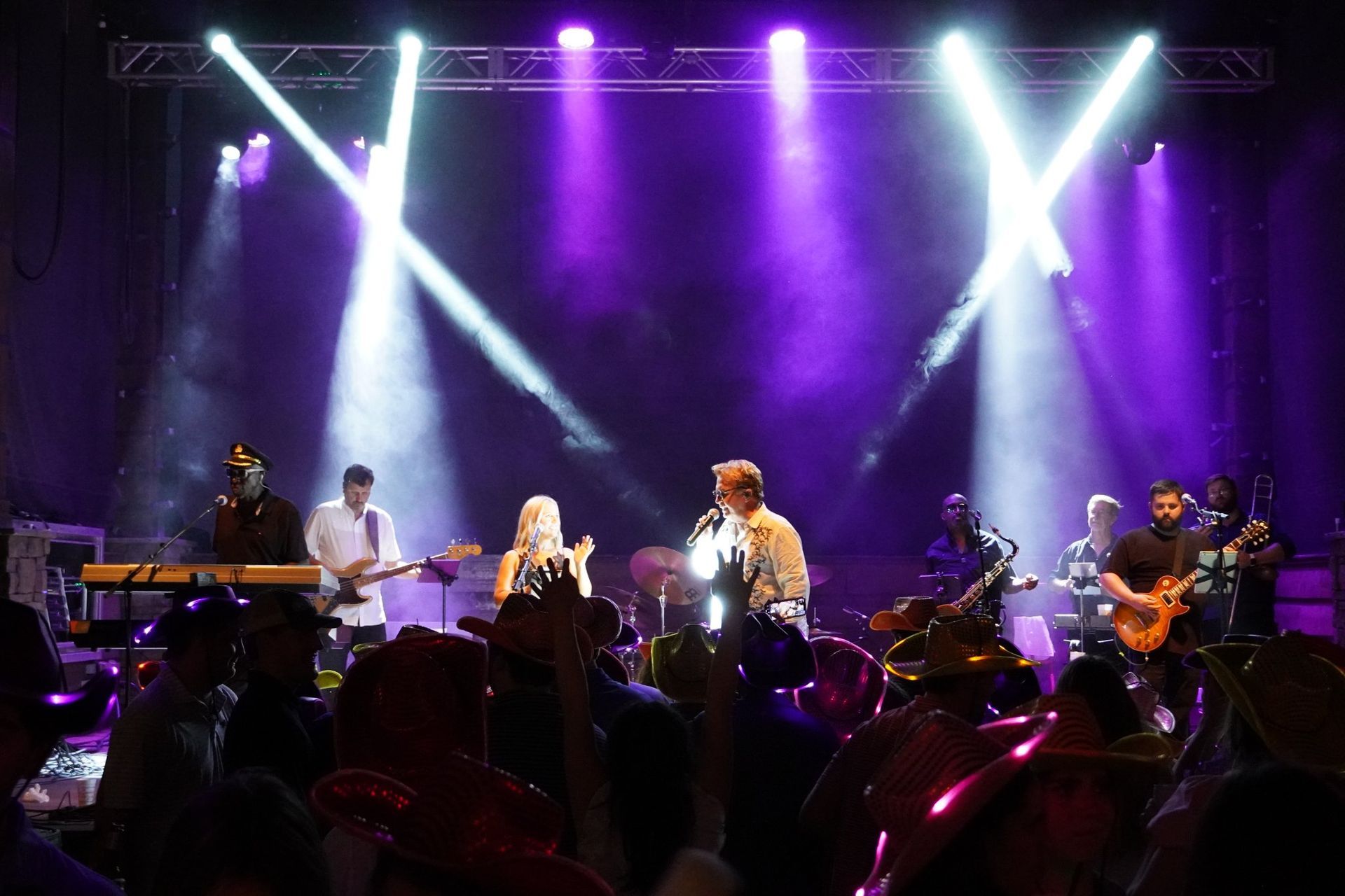 A band performs on a stage under purple lights, while audience members in the foreground have their hands raised.