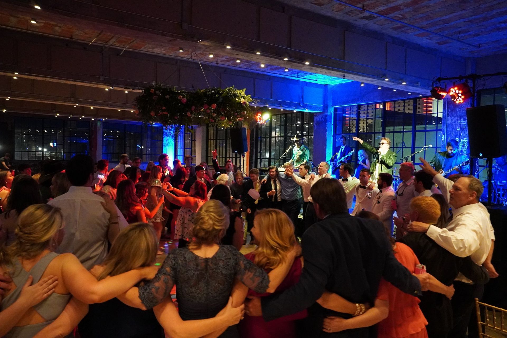A crowd at a wedding reception dances in a circle with arms linked under dim, blue-toned party lighting.
