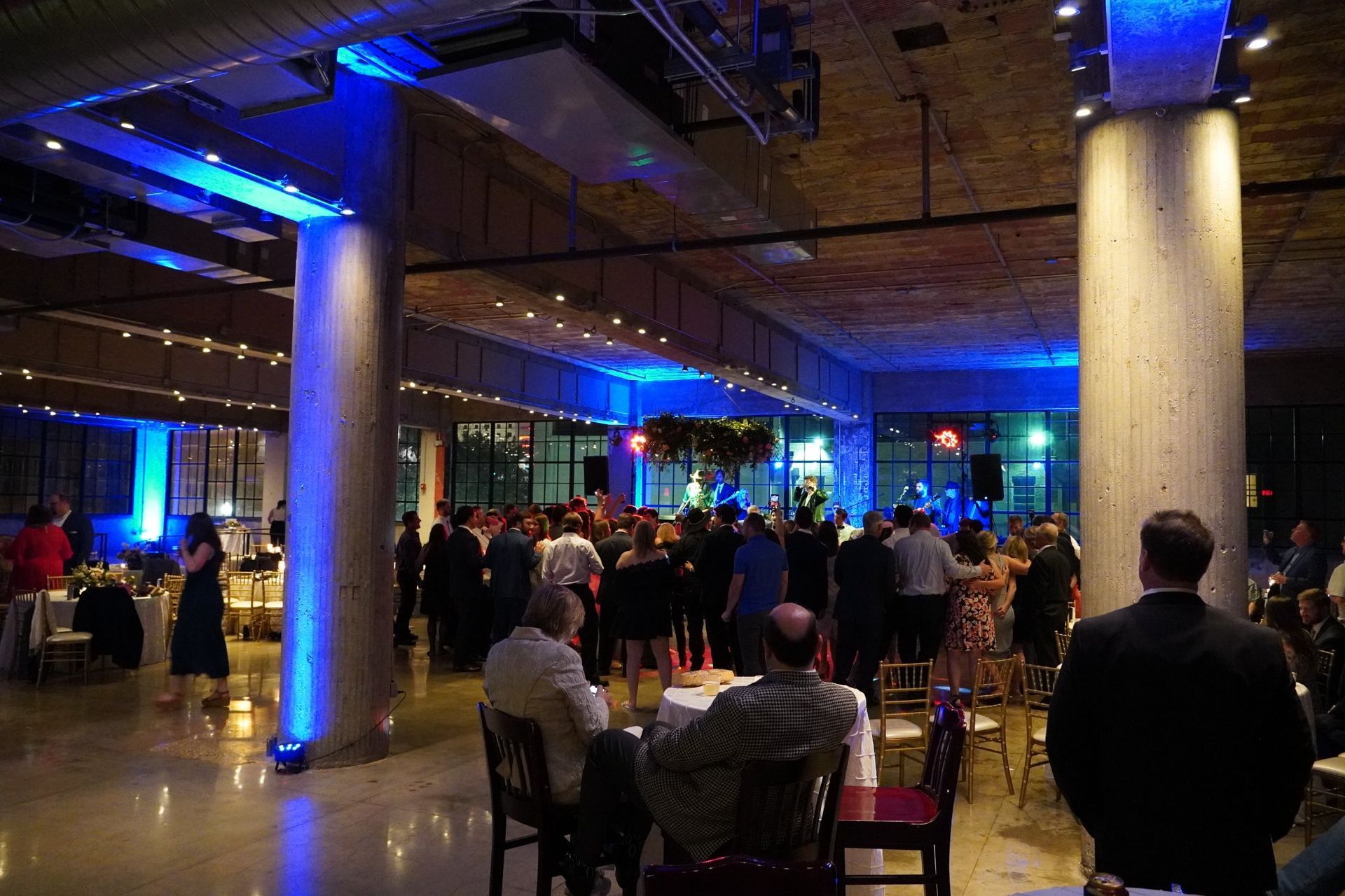 A dimly lit event space with concrete columns, blue ambient lighting, and guests mingling near a performance area.
