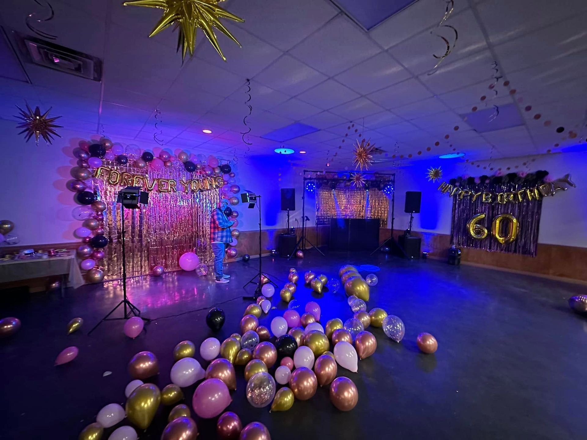 A room decorated for a 60th birthday party with gold fringe backdrops, balloons on the floor, and blue ambient lighting.