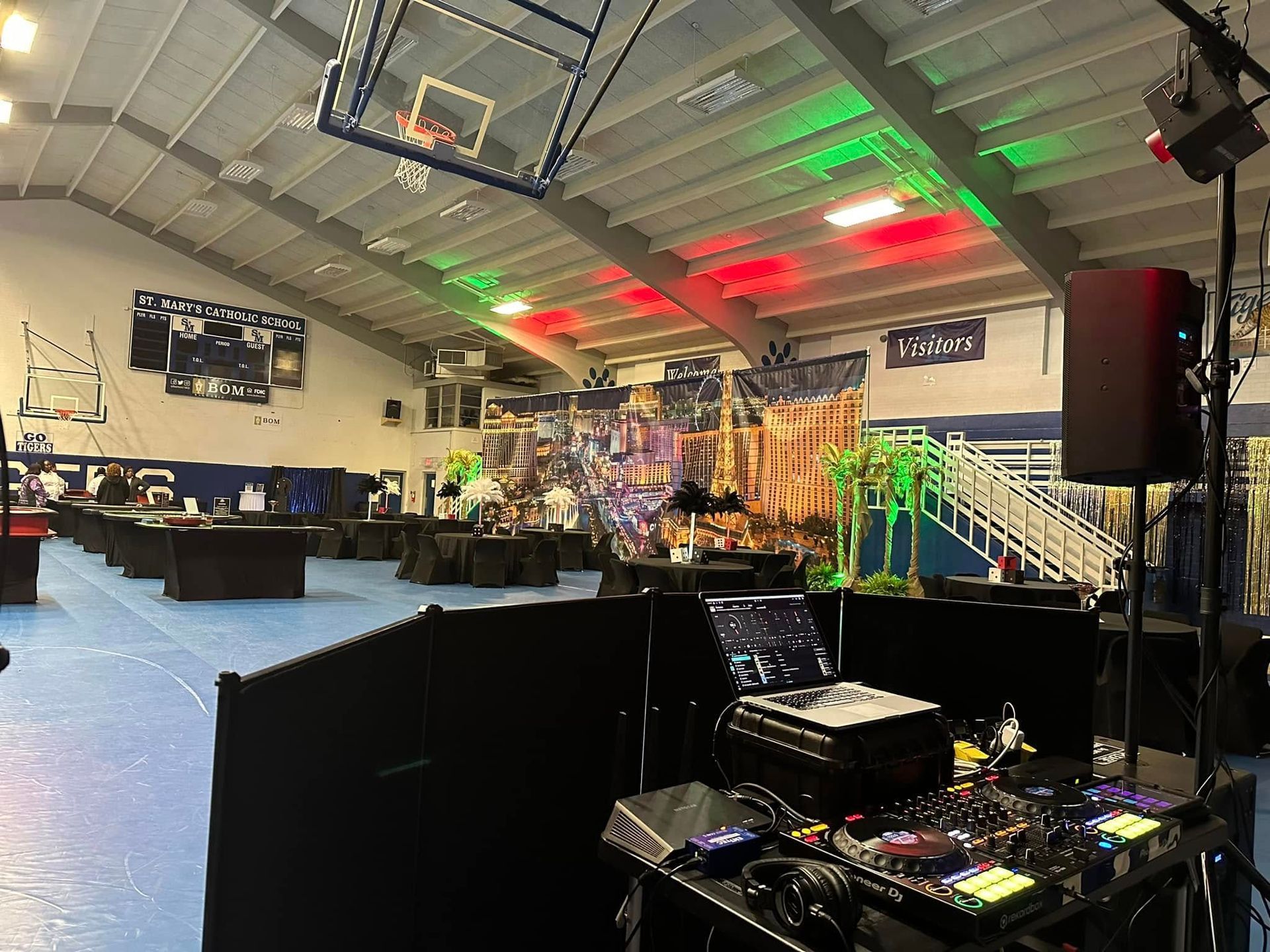 A DJ setup in the foreground overlooking an indoor gym decorated for an event with colored lighting and table seating.