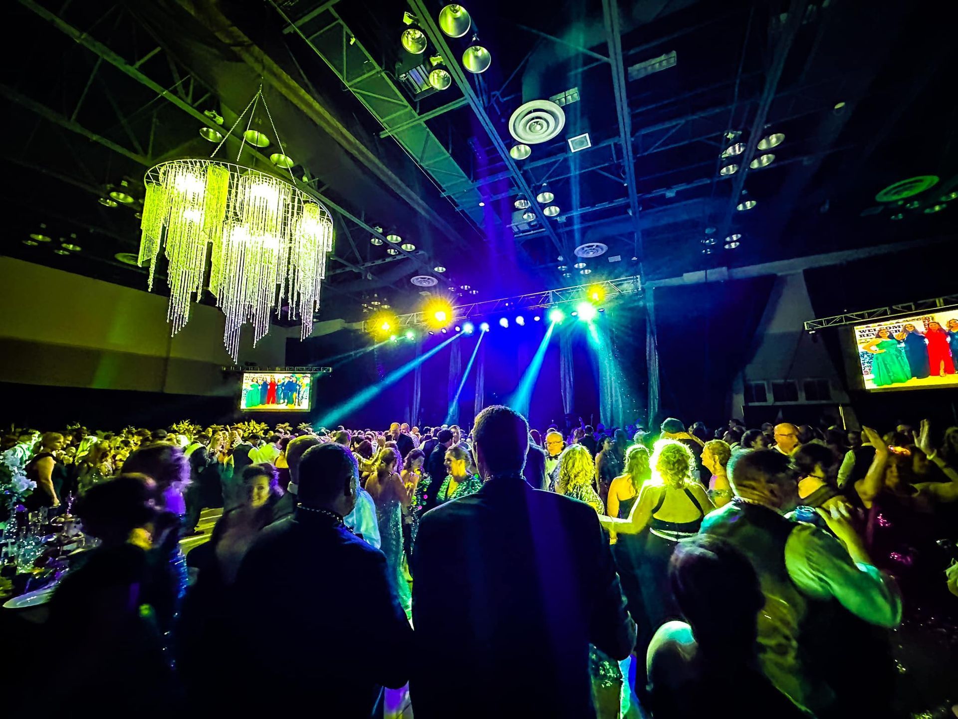 A crowded dance floor at a festive indoor event with bright stage lights, a large chandelier, and screen displays.