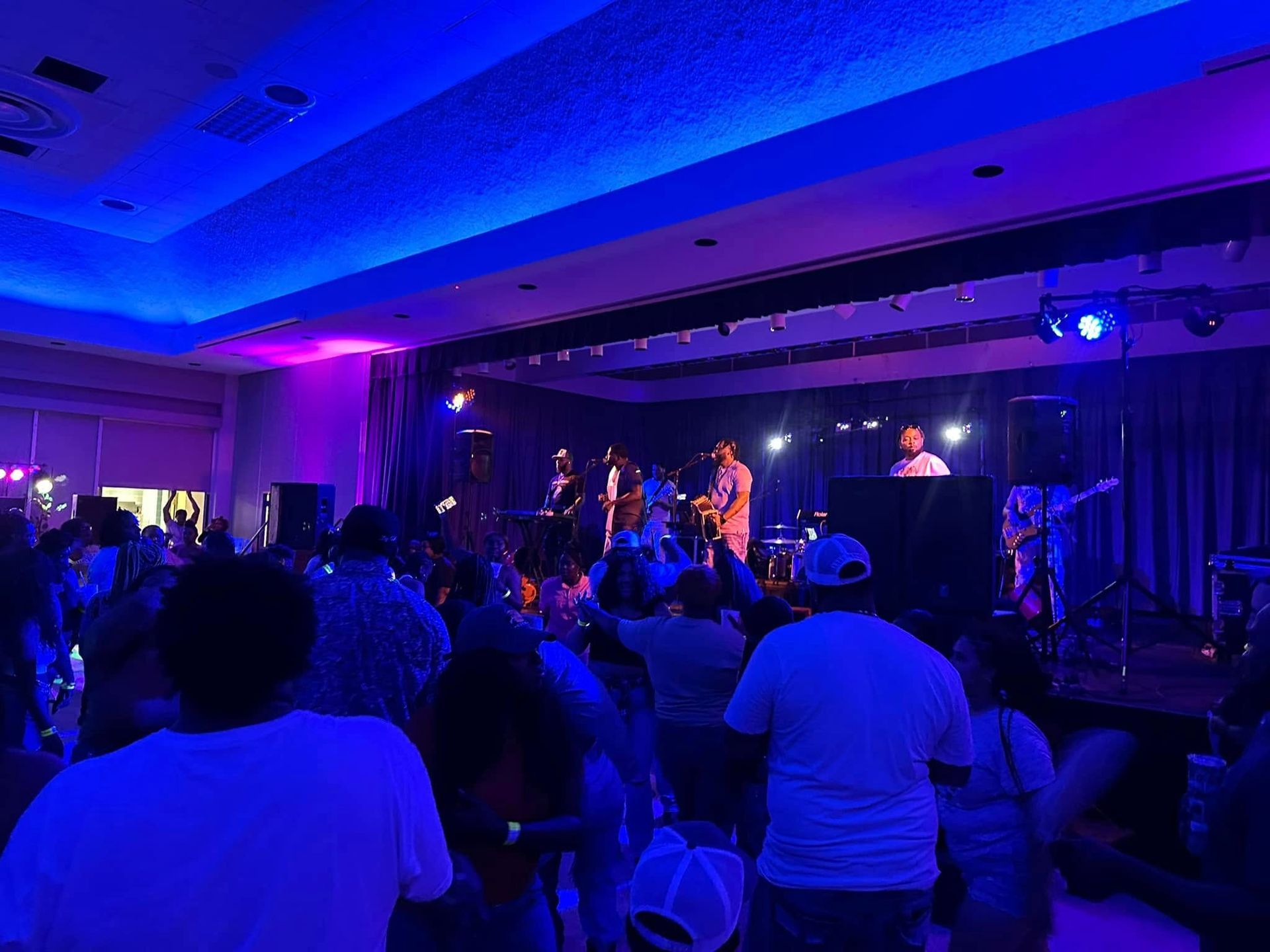 A band performs on a stage lit by blue and purple lights before a crowd of people in a dark event hall.