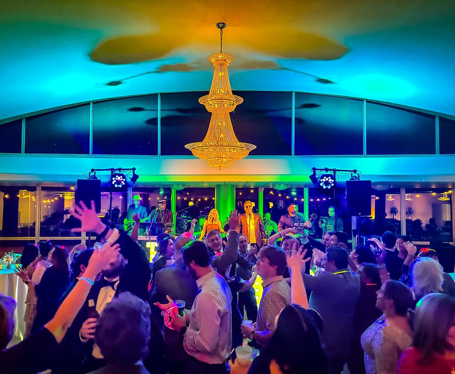 A crowd dances under a large, glowing chandelier in a venue illuminated by vibrant blue and green party lighting.