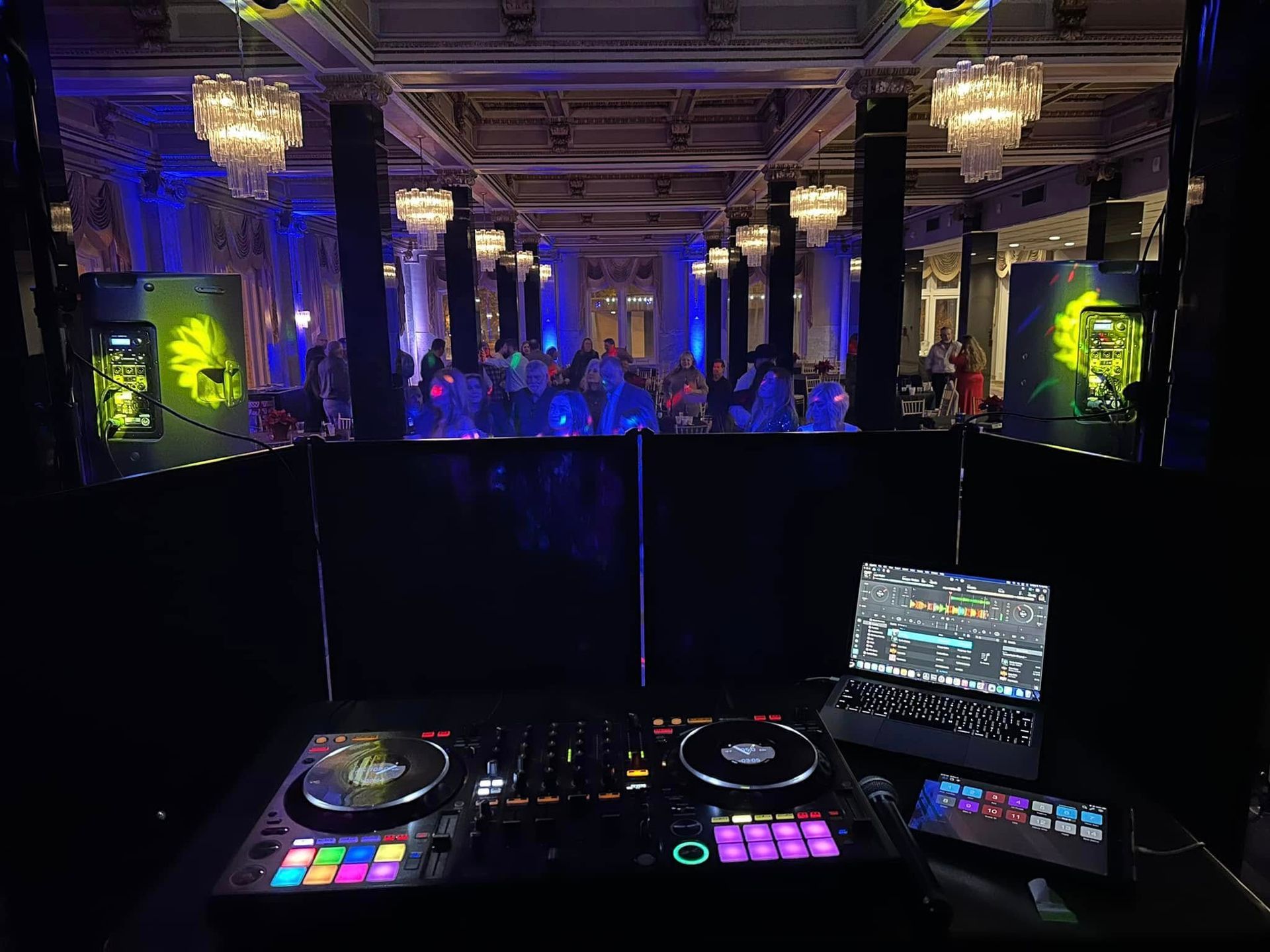 A DJ booth with mixing equipment, a laptop, and tablet overlooking a dimly lit, chandelier-adorned ballroom with dancers.