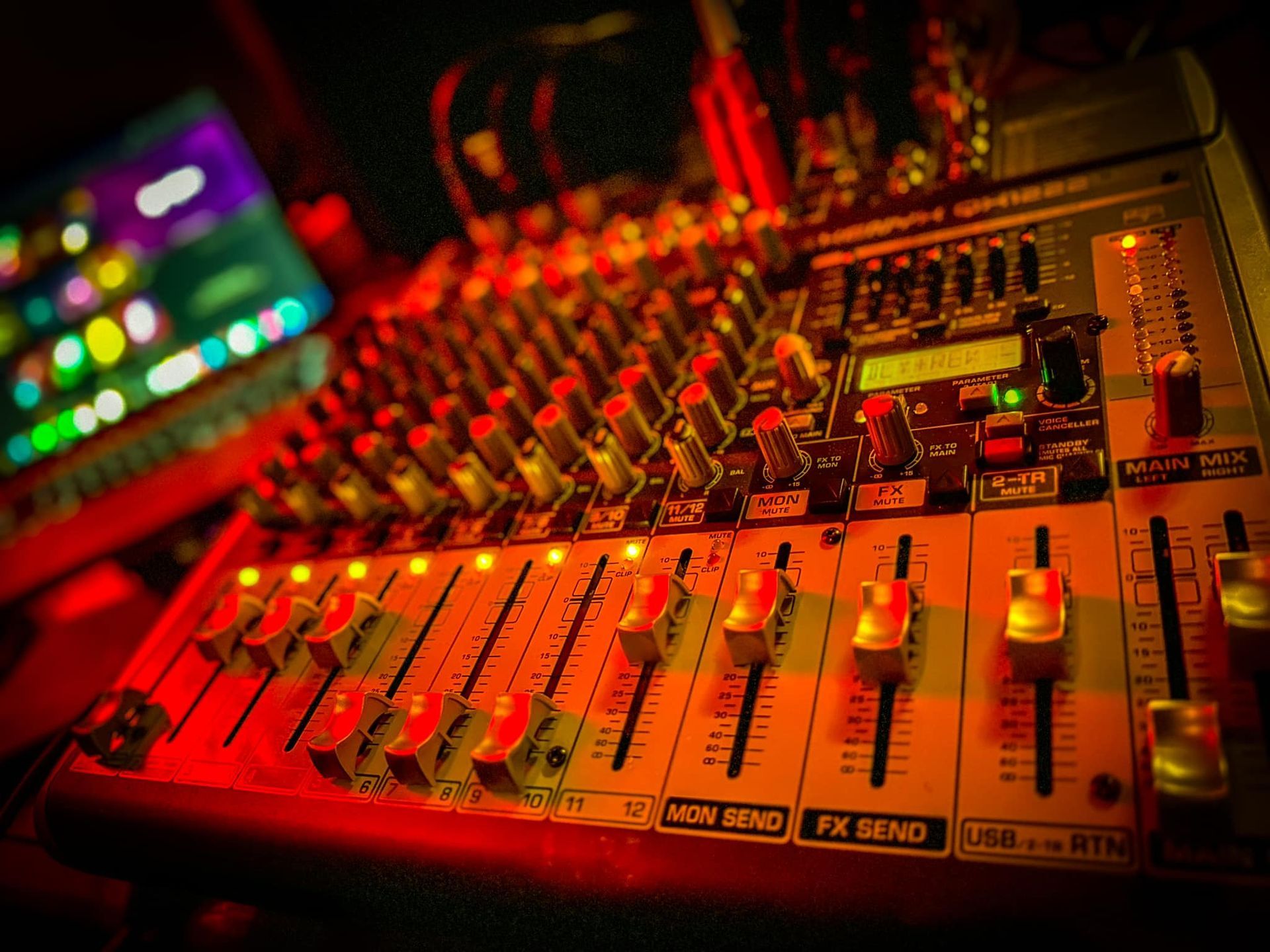 A close-up view of an audio mixing console illuminated by warm red lighting, with a blurred background.