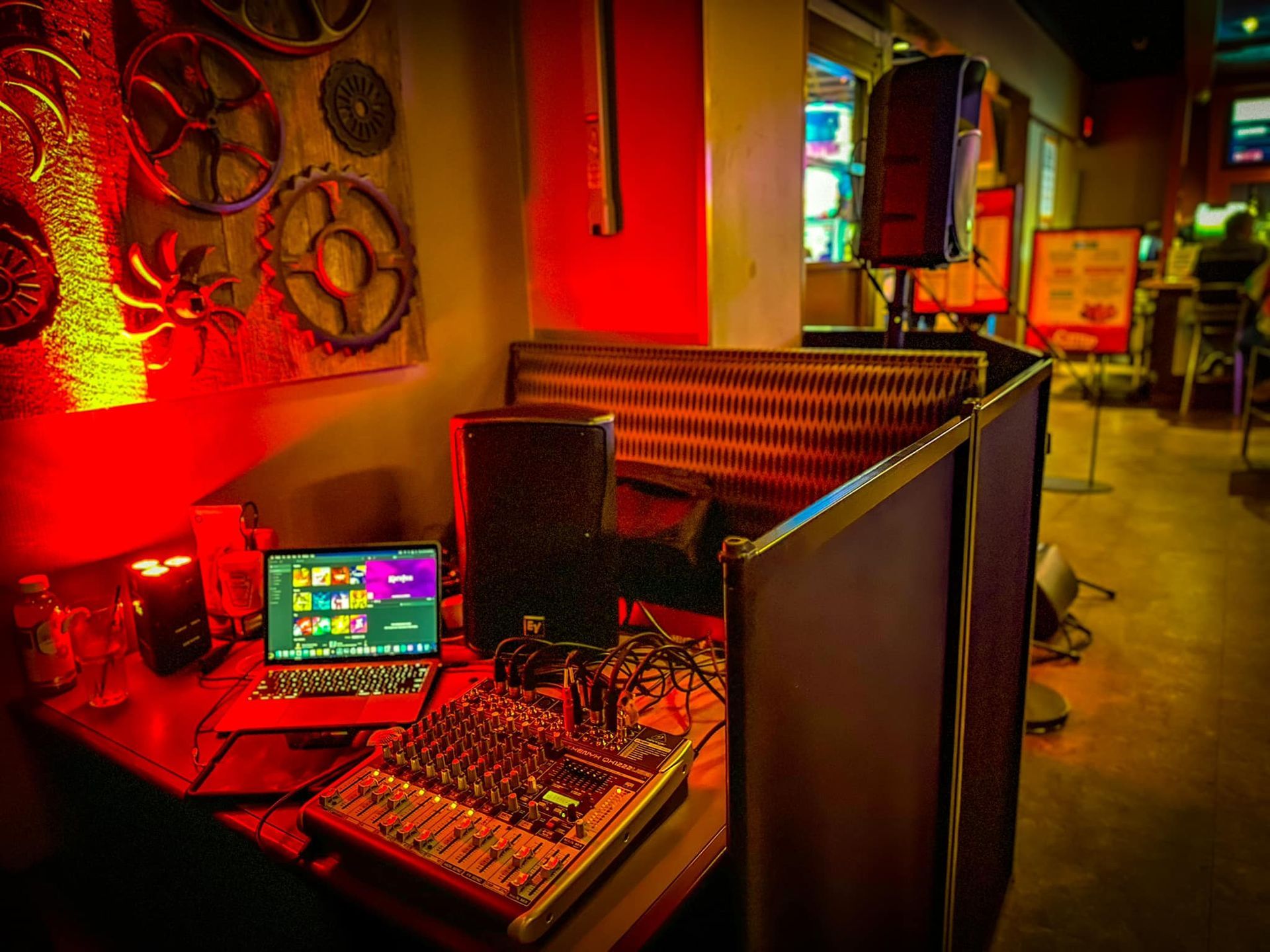 A DJ booth with a laptop, audio mixer, and speakers in a dimly lit, red-lit venue decorated with industrial wall gears.