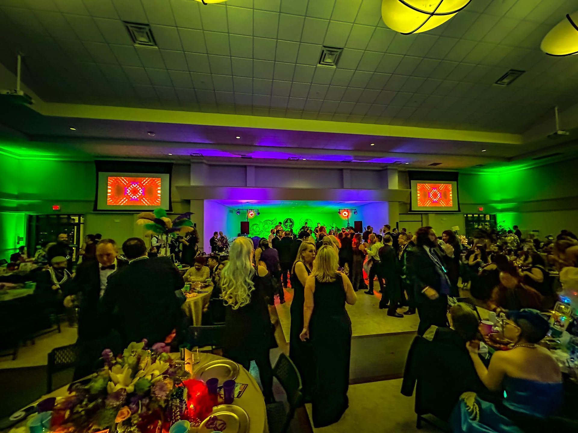 People in formal wear attend a lively event in a large, dimly lit hall decorated with green and purple ambient lighting.