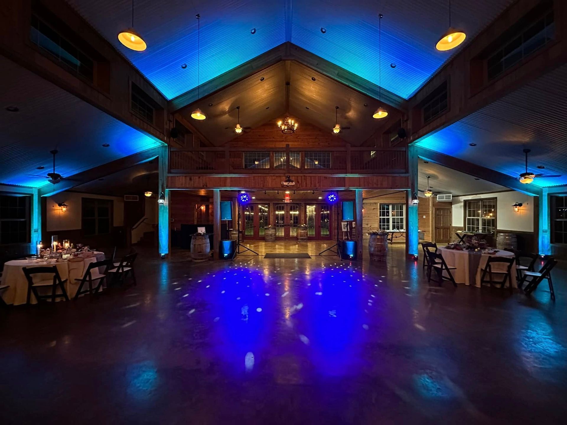 A wide view of a rustic event venue featuring wood beams, tables, and dramatic blue mood lighting on the dance floor.
