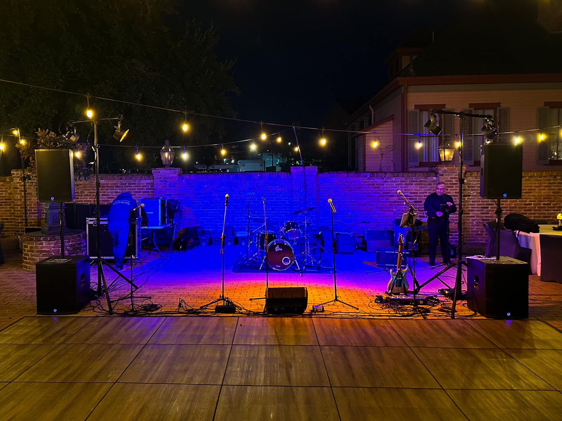 An outdoor stage at night with a drum kit, speakers, and string lights, illuminated by blue and orange stage lighting.