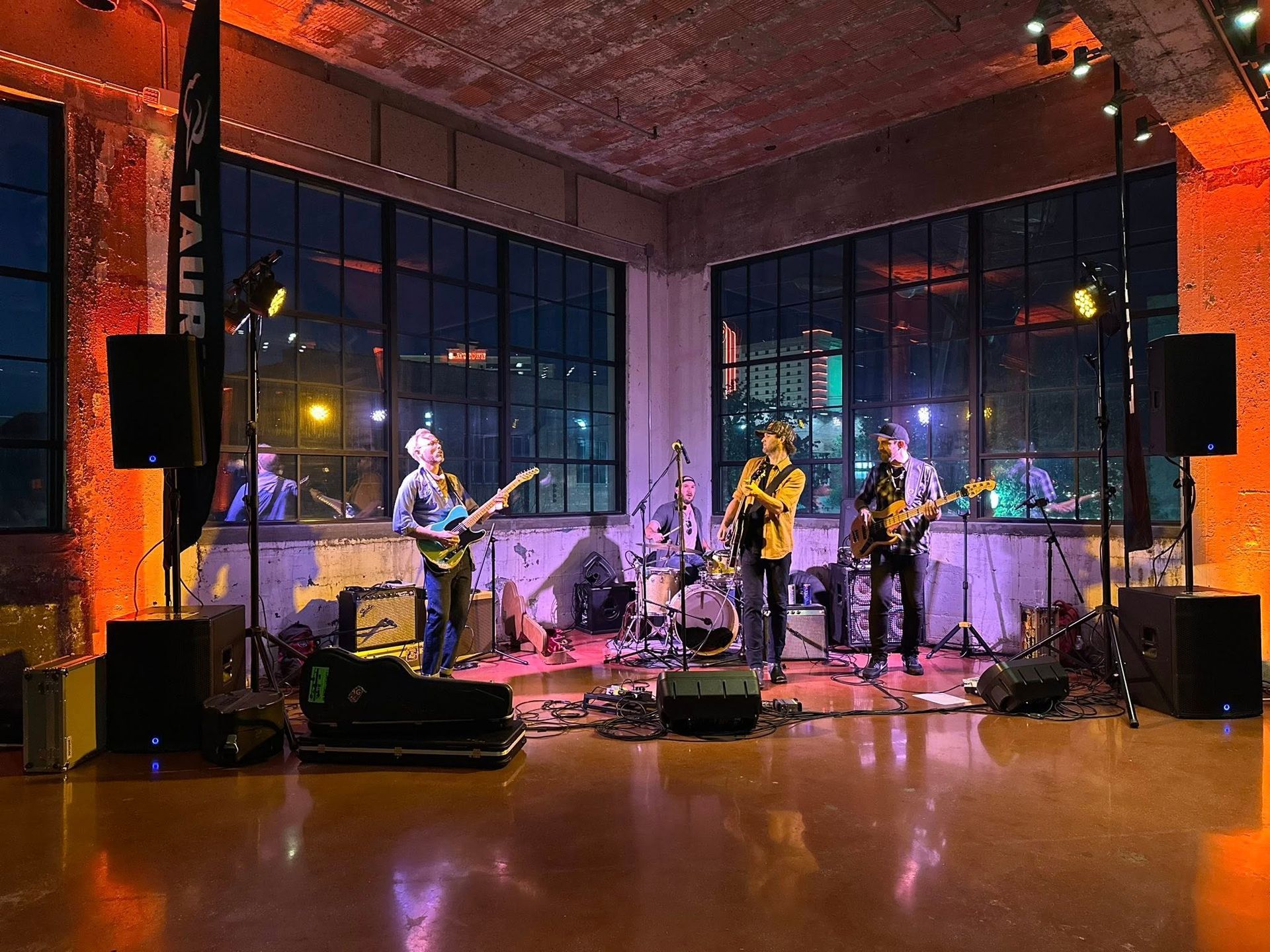 A four-piece band performs on a stage with large windows, warm lighting, and music equipment in an industrial-style room.