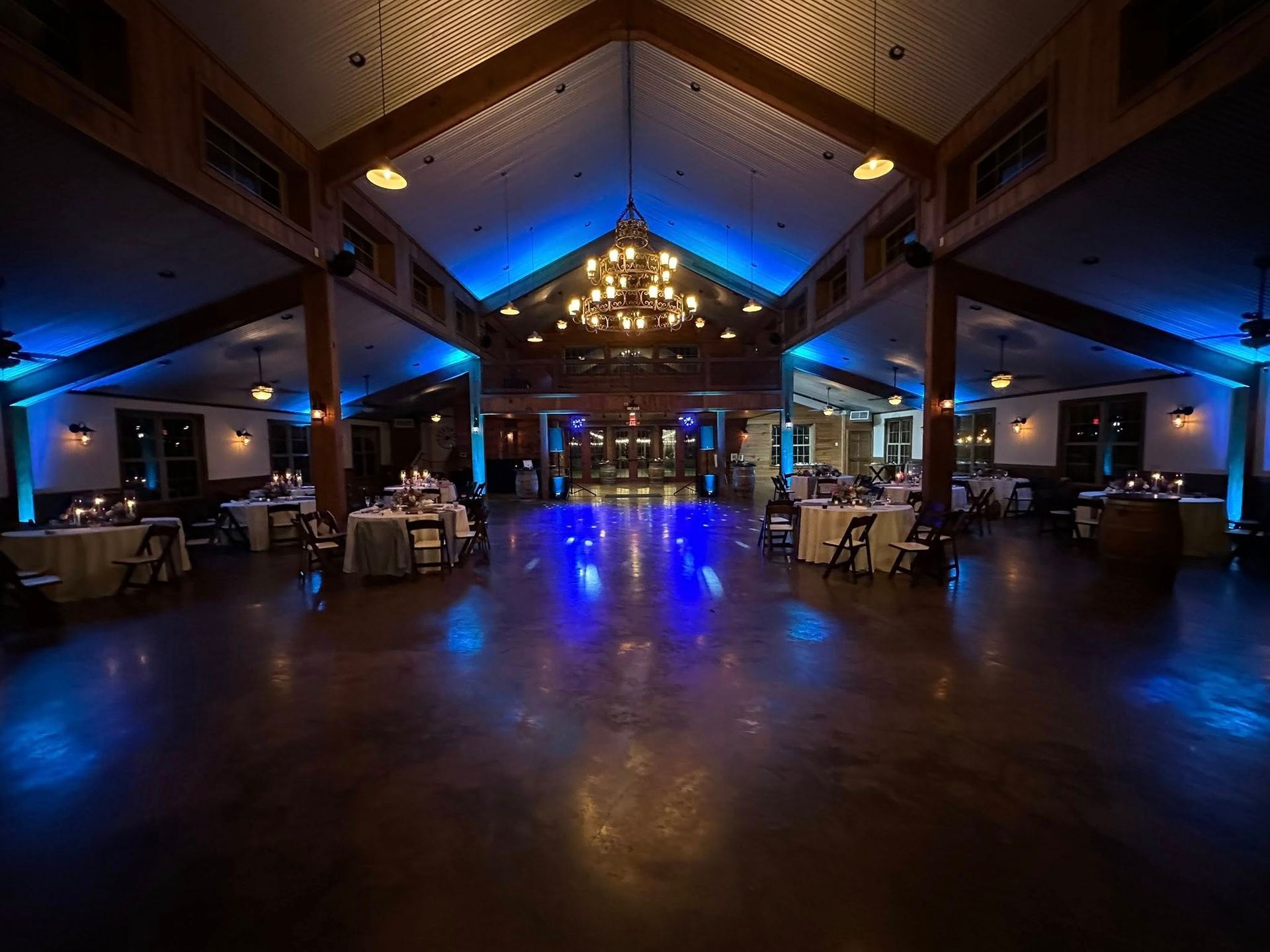 A spacious event hall with vaulted wooden ceilings, a central chandelier, and blue accent lighting over round tables.