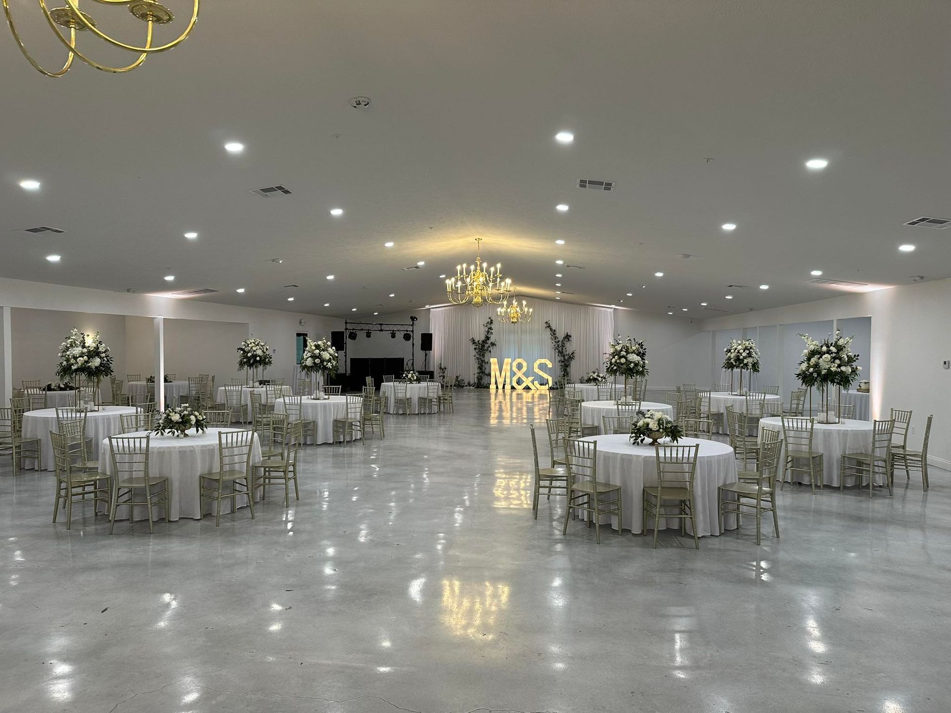 A spacious, white-walled event hall featuring circular tables with floral centerpieces and illuminated 