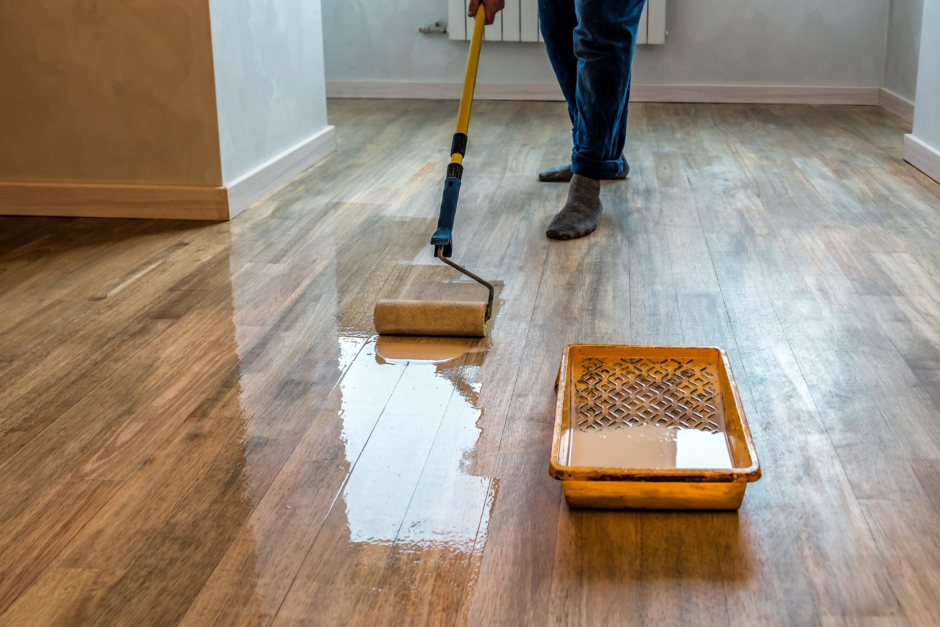 Person applying sealant to a wooden floor with a paint roller and tray indoors