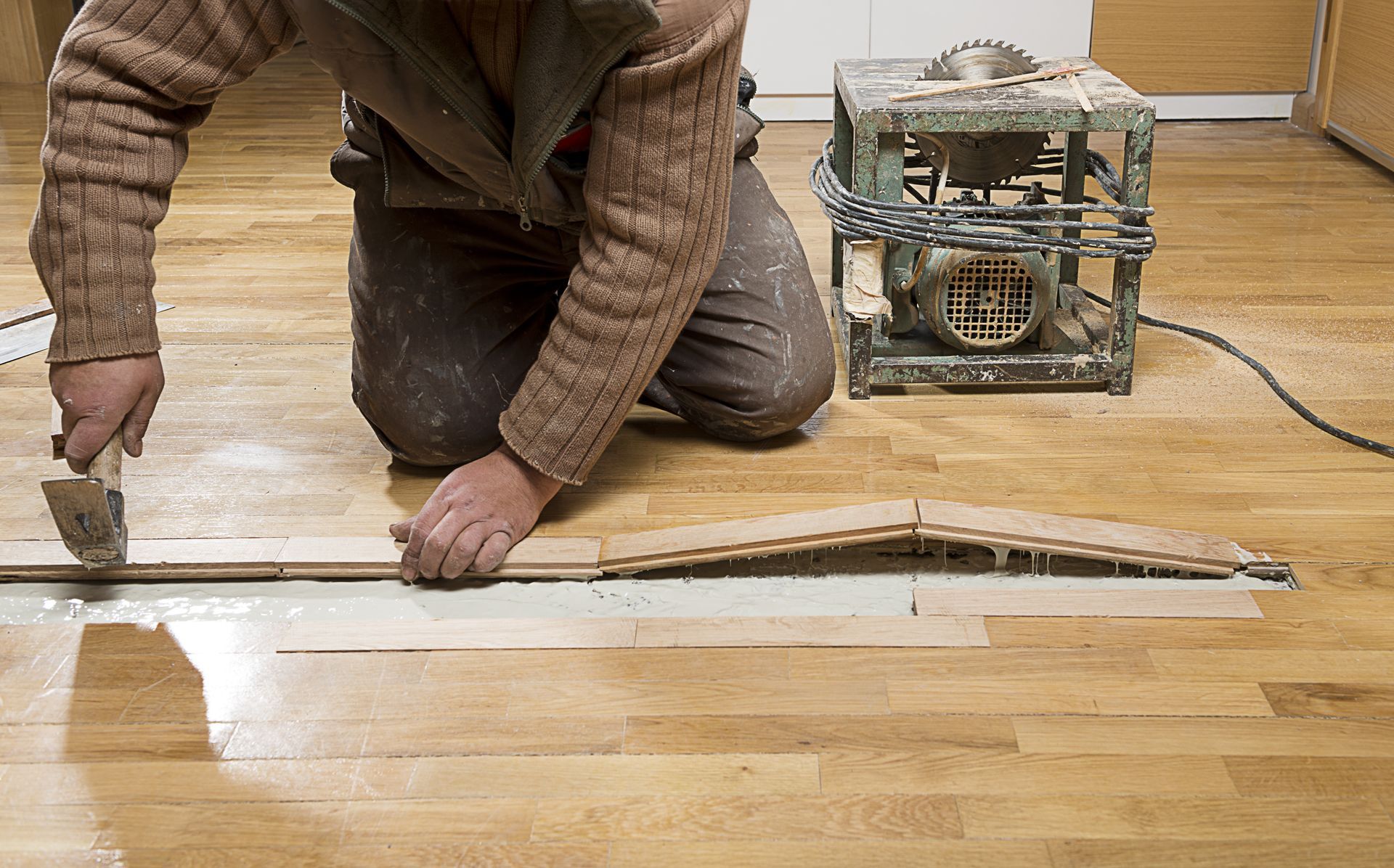 Person kneeling on wooden floor, applying glue with trowel, near a circular saw