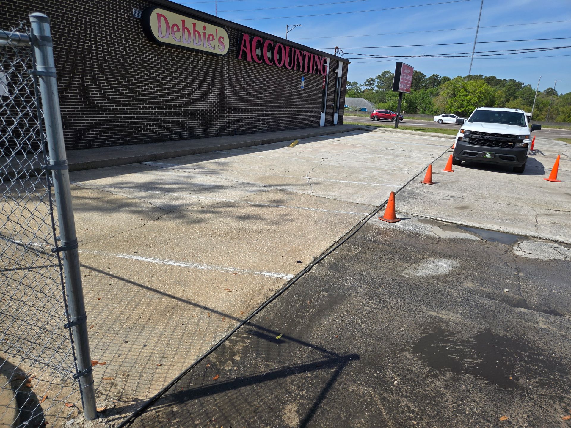 A light-colored parking area bordered by a chain-link fence and a building. A white truck and orange cones are visible.