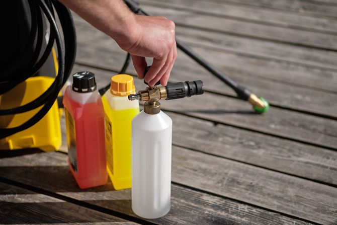 Person adjusting a foam cannon attached to a bottle, next to cleaning solution bottles and pressure washer equipment on wooden deck.